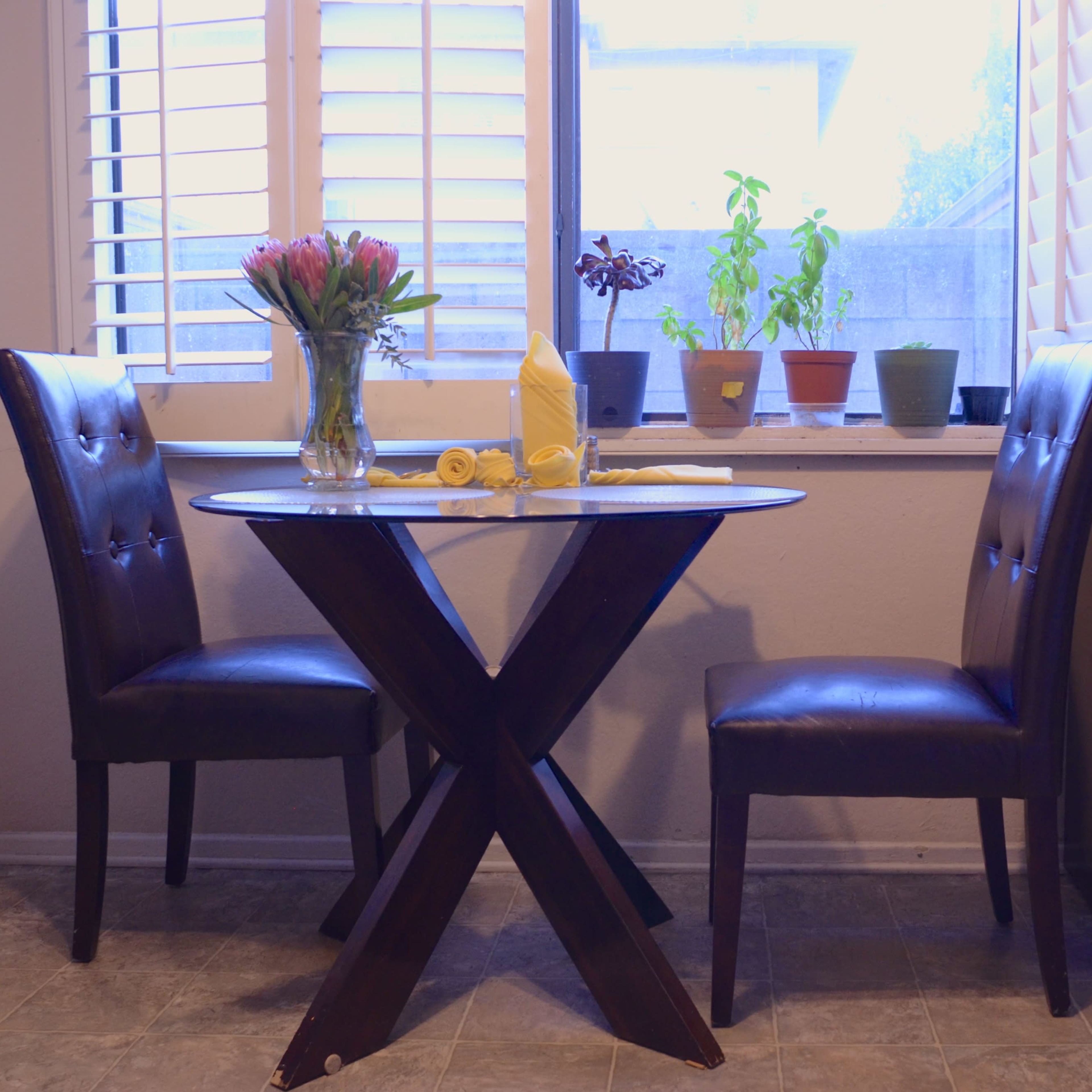 A small kitchen corner features a round table with two chairs, a vase of flowers, and several potted plants near a window.
