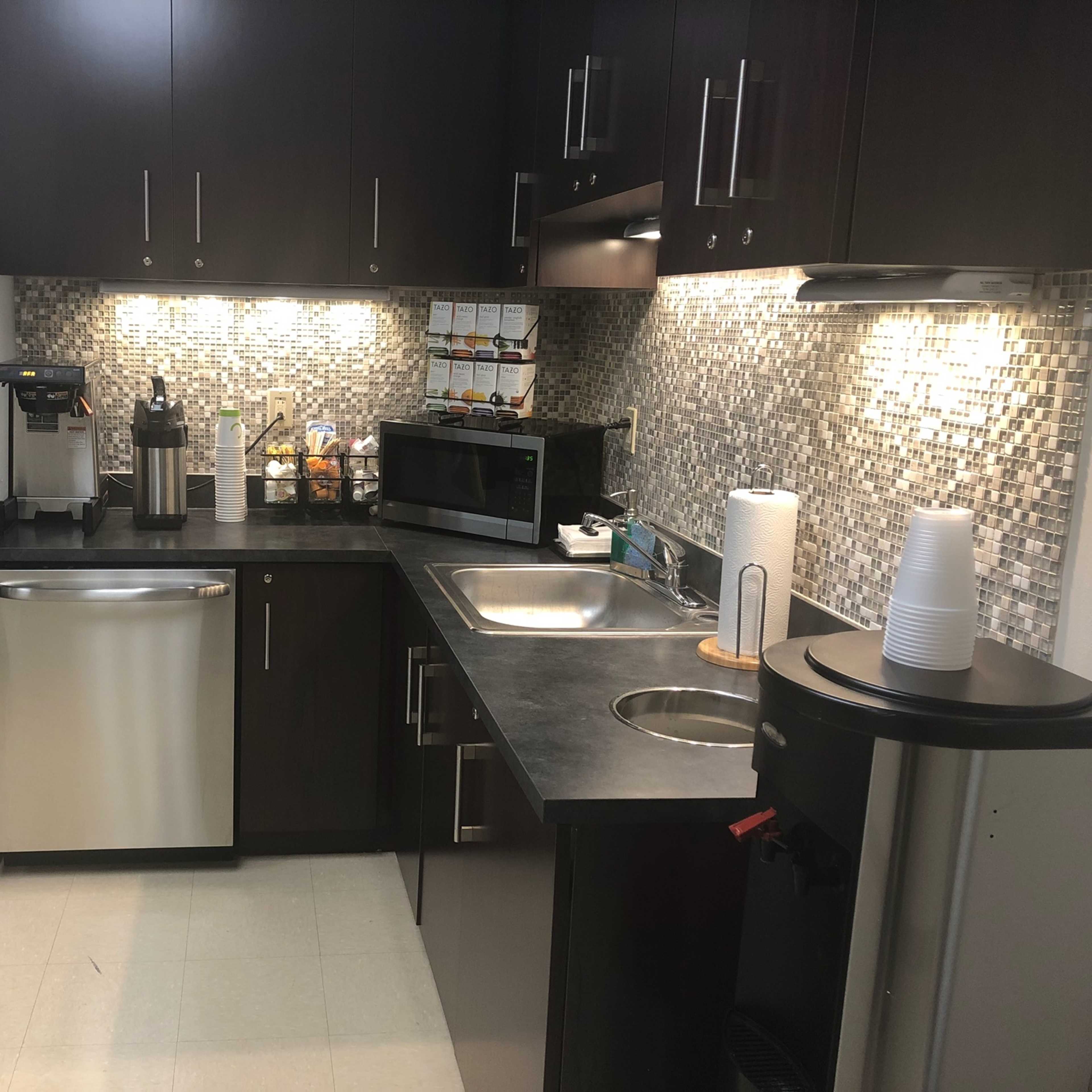 A modern kitchen with stainless steel appliances, including a refrigerator and dishwasher, a microwave, and a coffee machine, set against a tiled backsplash.