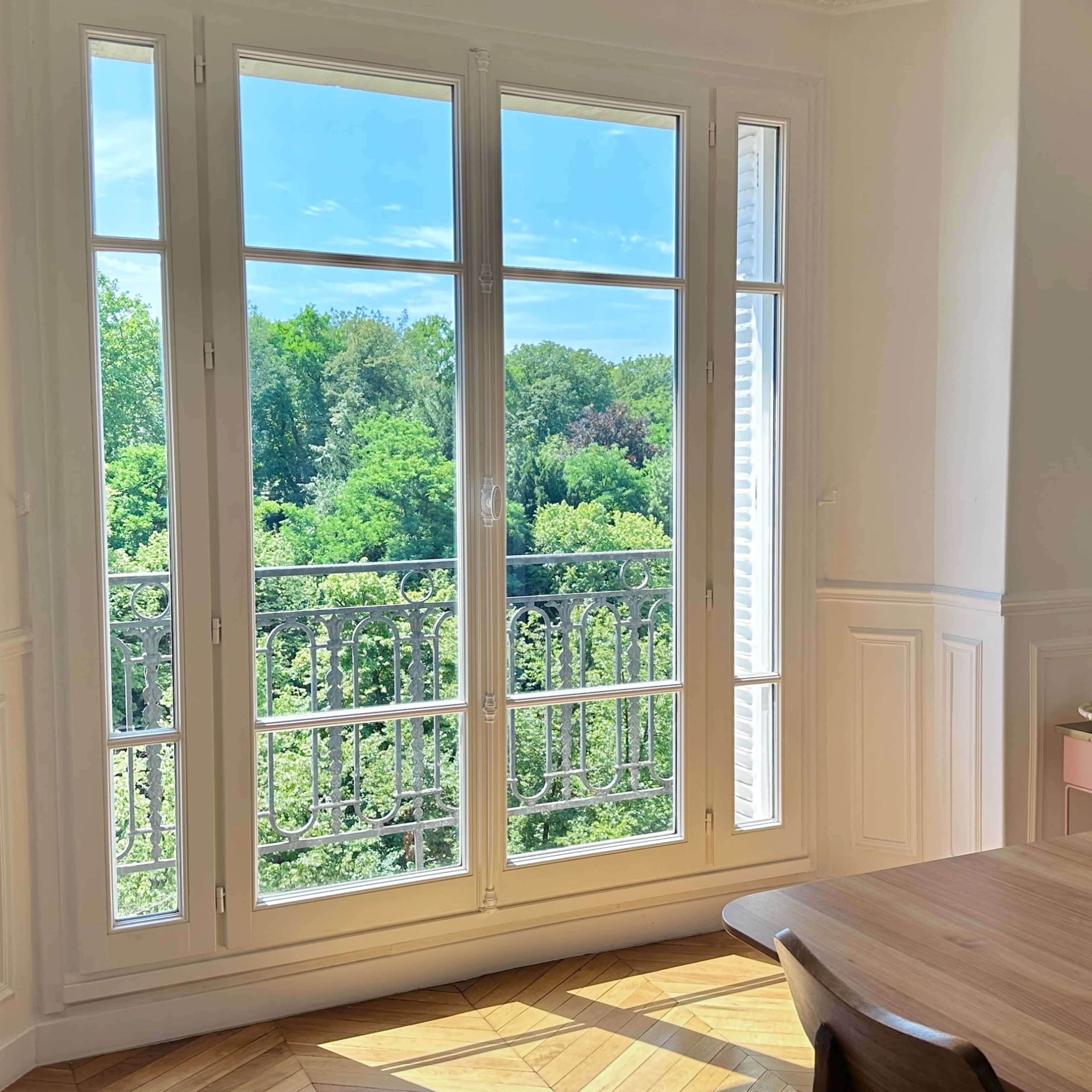 A bright room features a large window opening to a view of lush greenery outside, with a dining table set nearby.