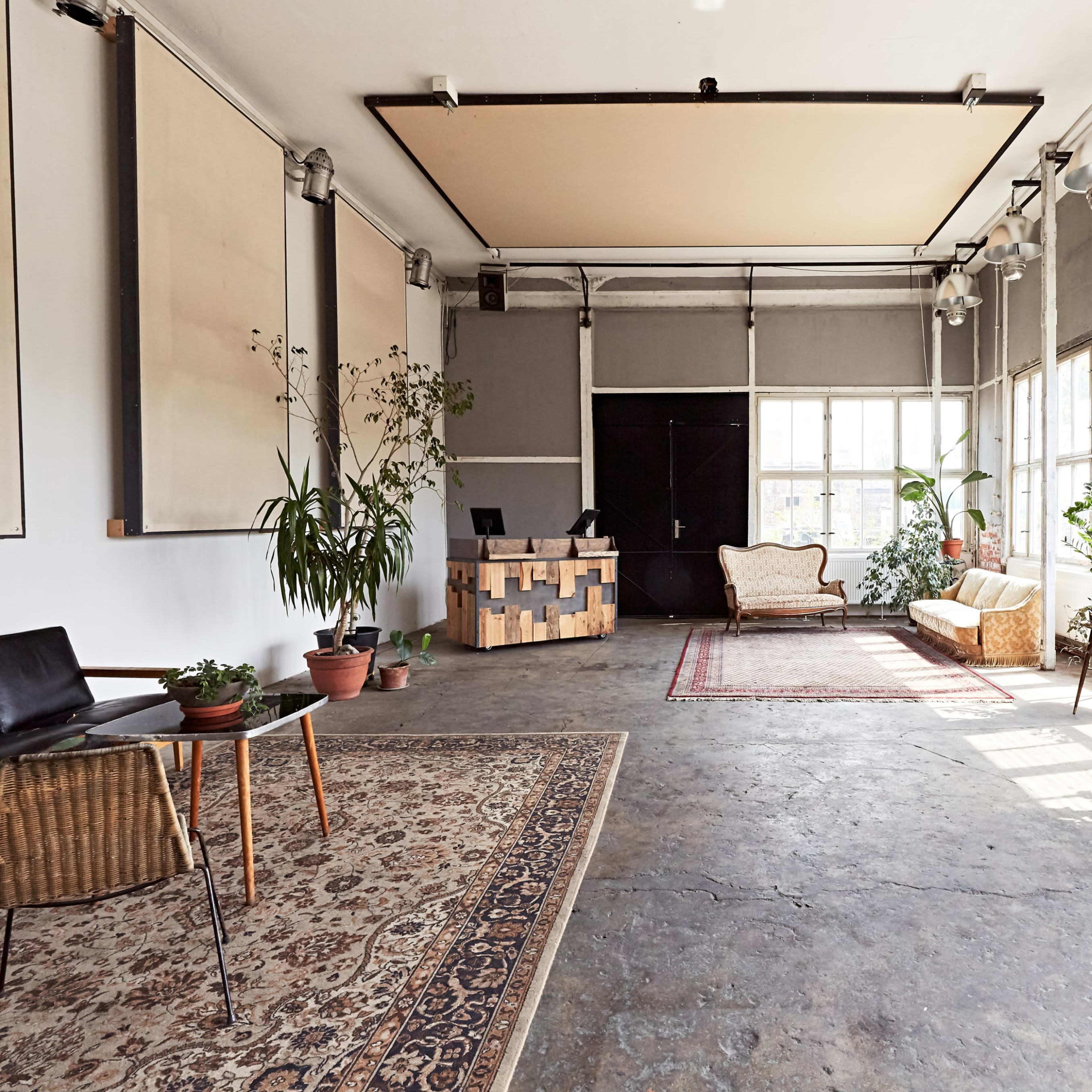 A spacious, well-lit interior features large windows, plants, a reception desk, and a seating area with rugs on a concrete floor.