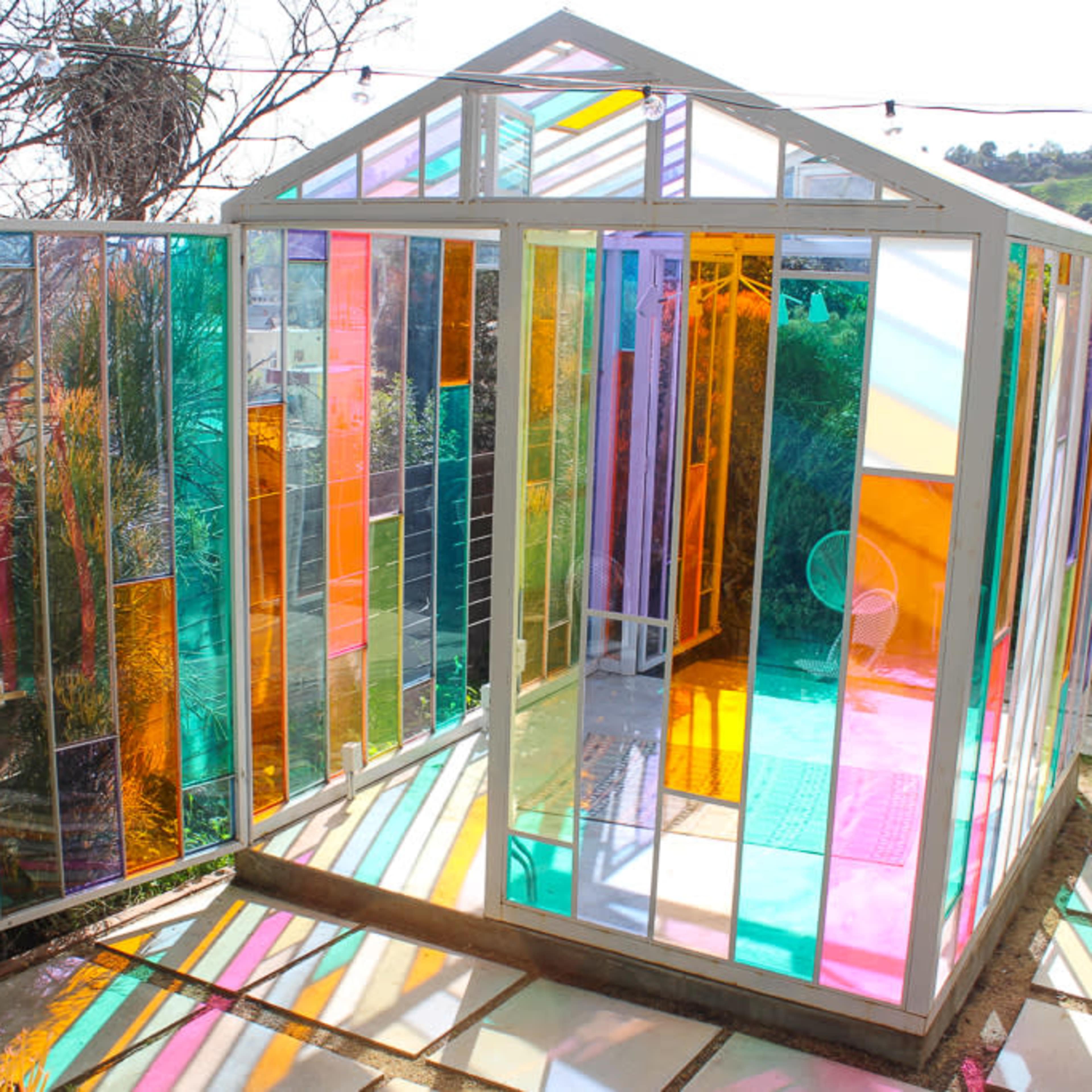 A colorful glass greenhouse with multicolored panels and an open doorway, situated in a garden with plants and outdoor tiles.