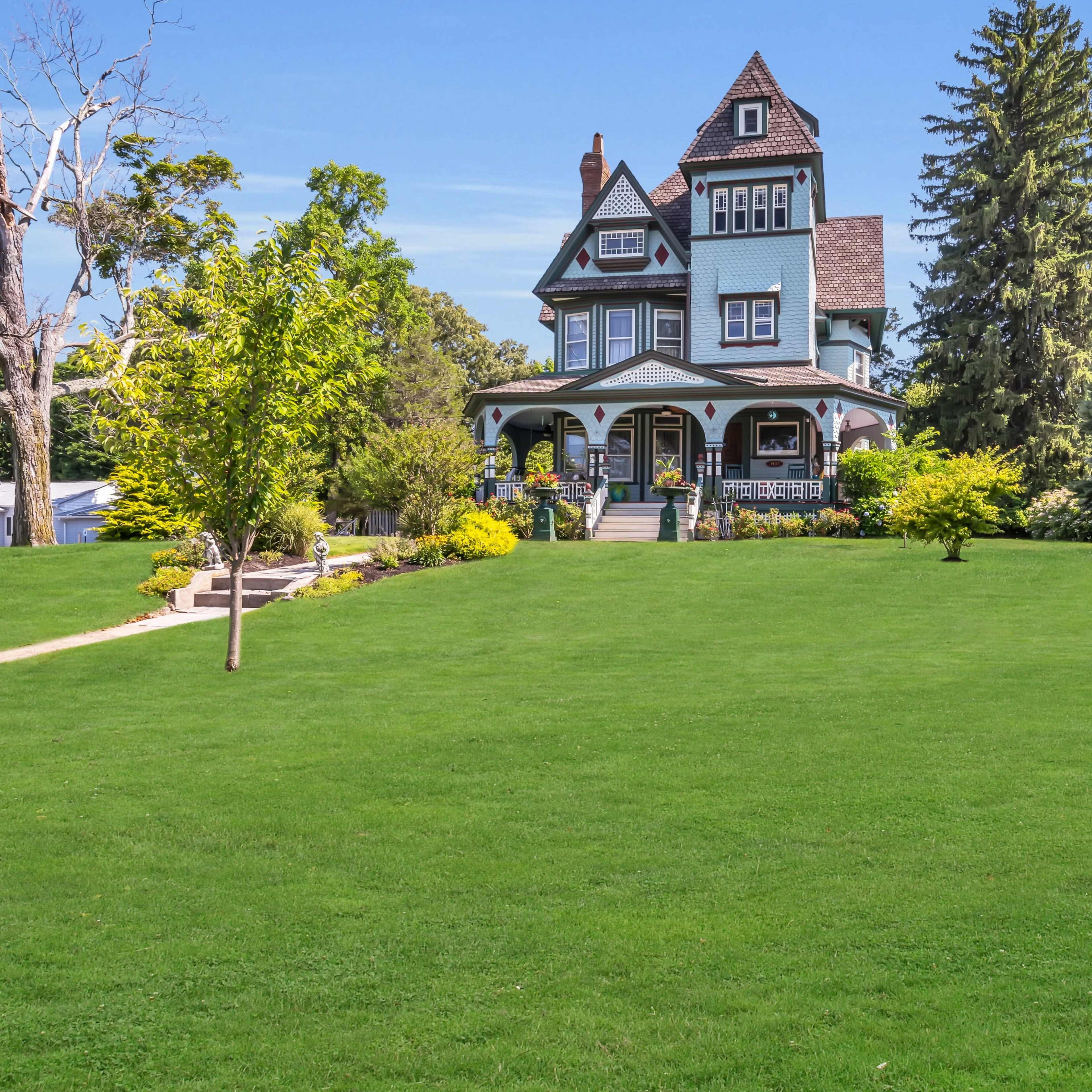 A large, multi-story Victorian-style house with a turret sits surrounded by a lush green lawn and several trees.