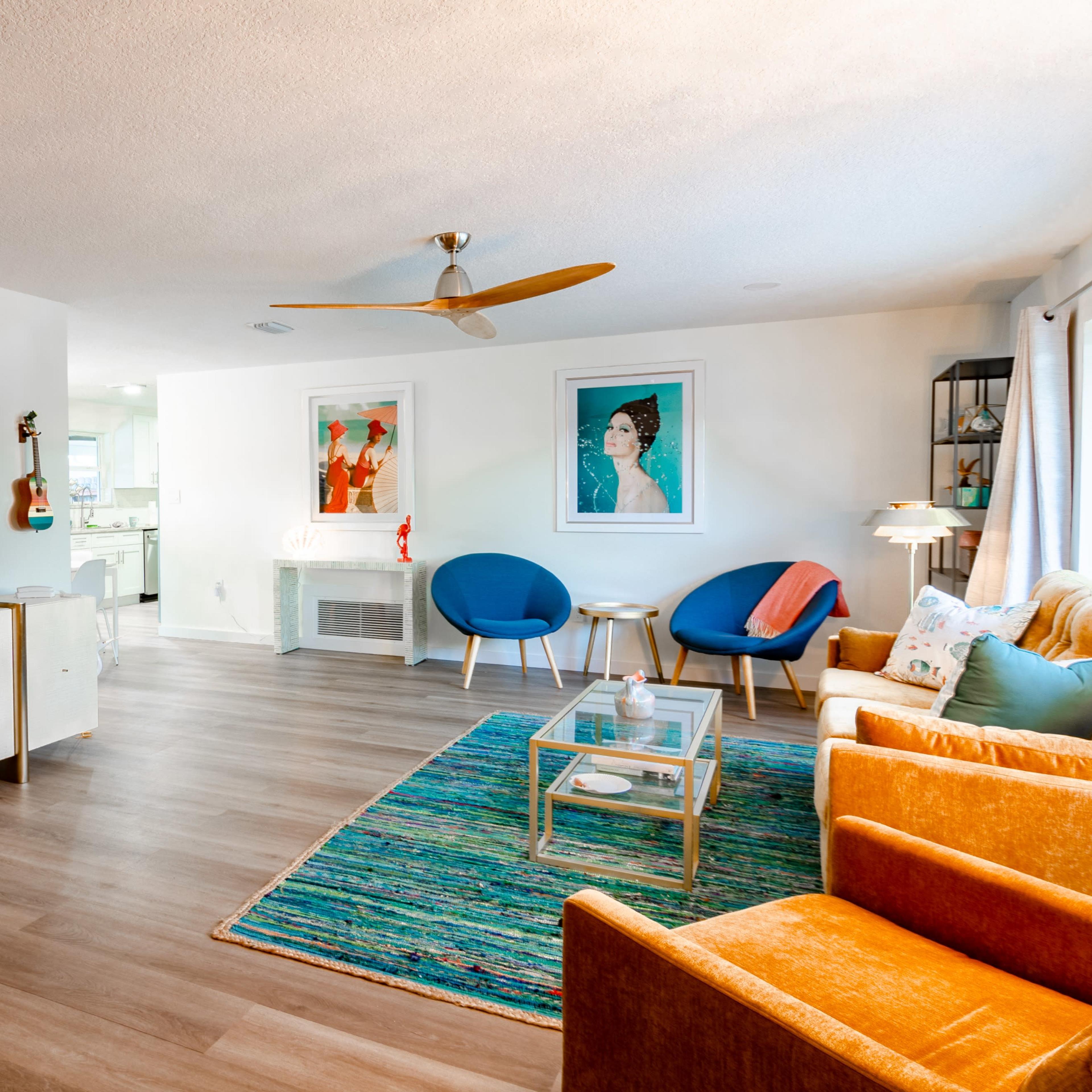 A modern living room featuring a large television mounted on the wall, a glass coffee table, colorful chairs, and a bright area rug.