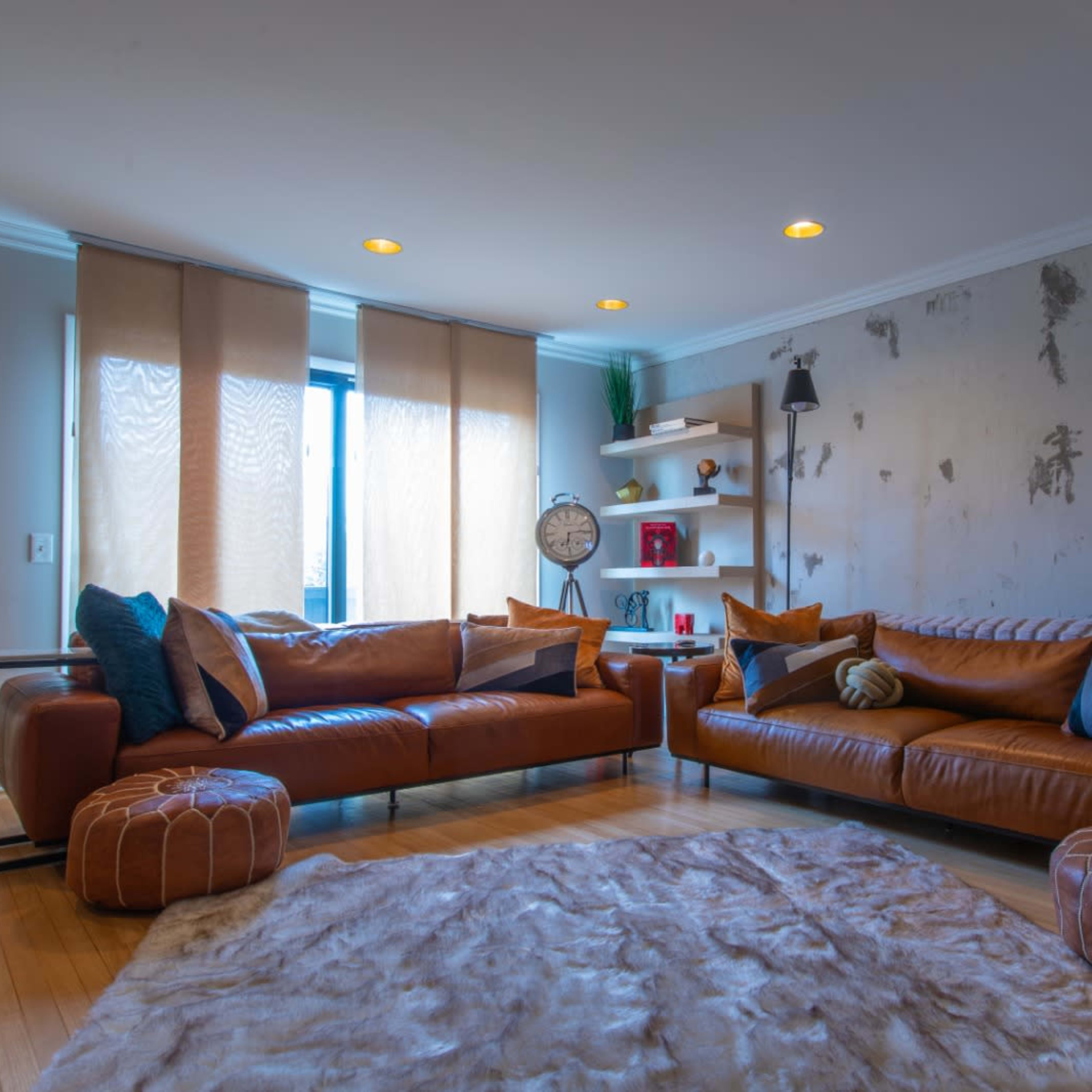 A modern living room features two leather sofas arranged opposite each other, with decorative pillows, a plush rug, wall shelves, and natural light from large windows.