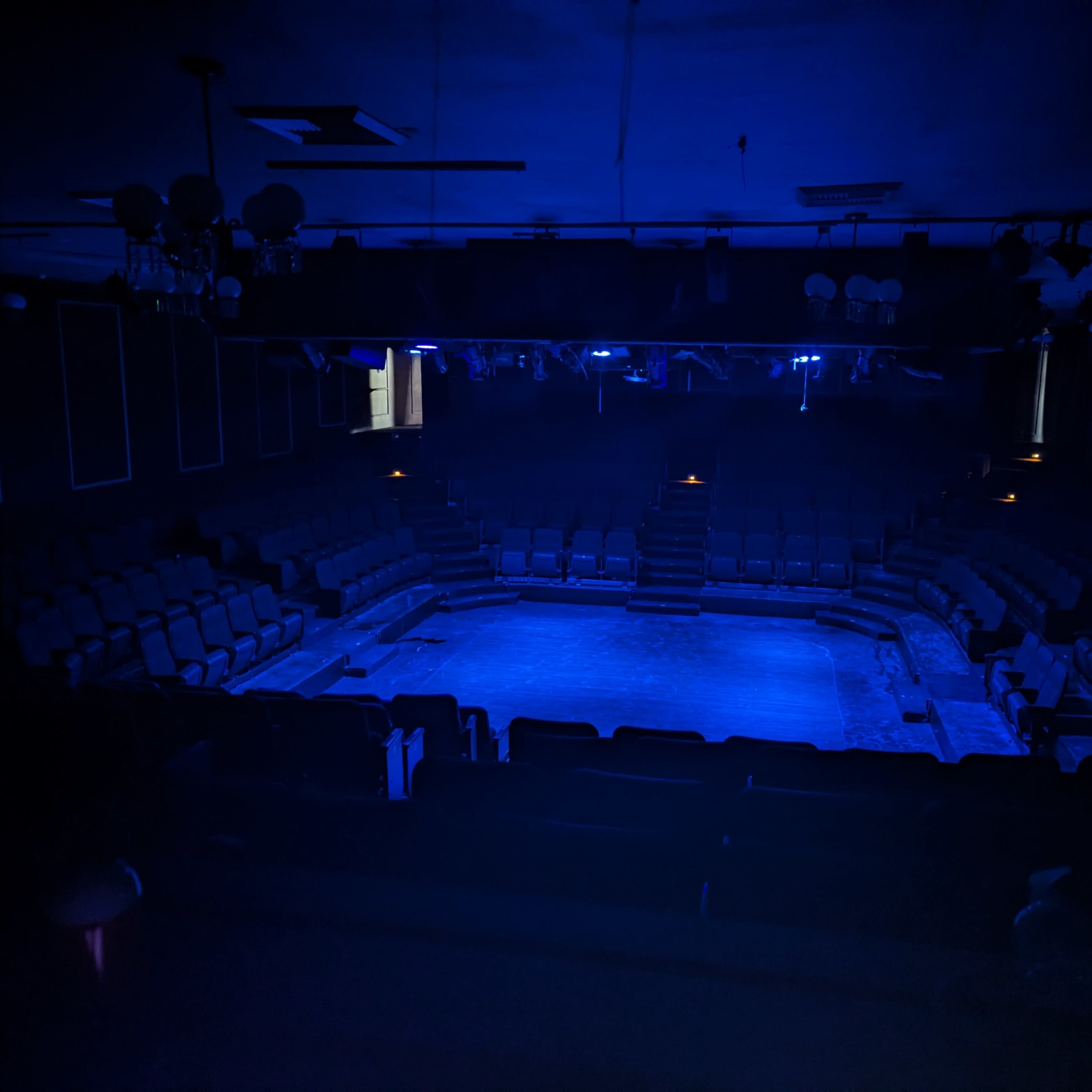A dimly lit theater with blue lighting and empty seating surrounding a central stage area.