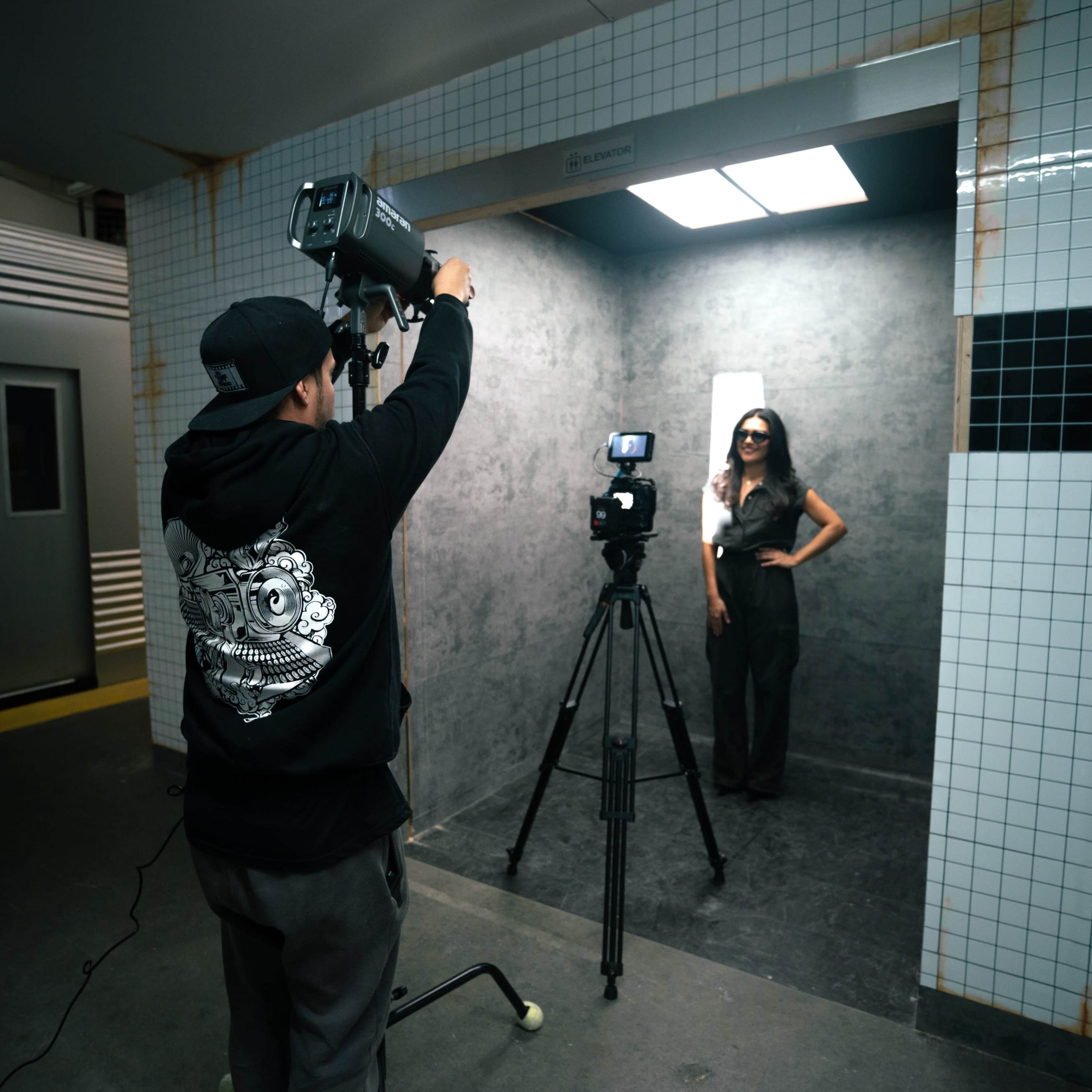 A photographer sets up a lighting rig while a model poses in a photo shoot against a tiled backdrop in a subway station.