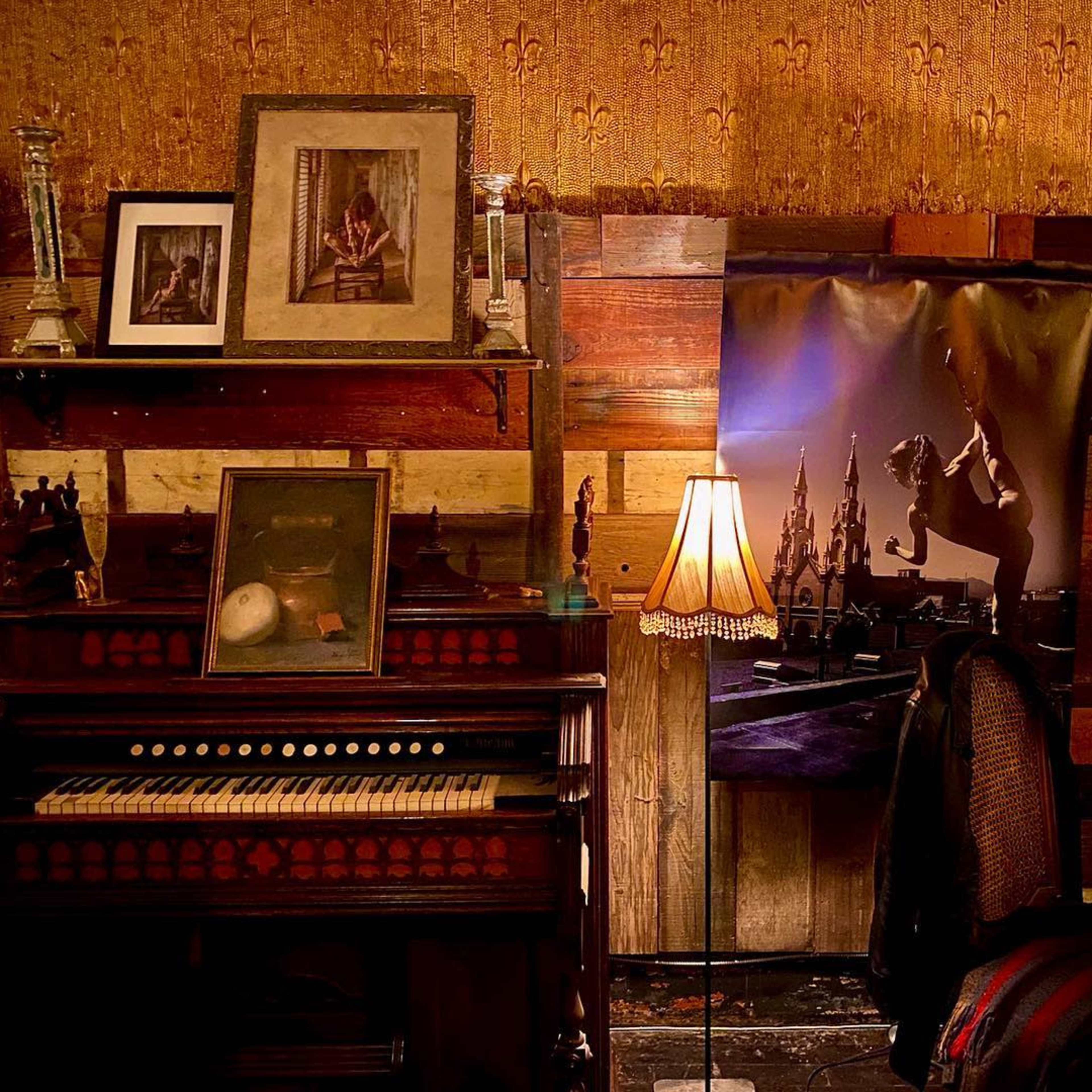 The scene features an antique piano surrounded by framed pictures on a wooden wall, with a decorative lamp casting light nearby.