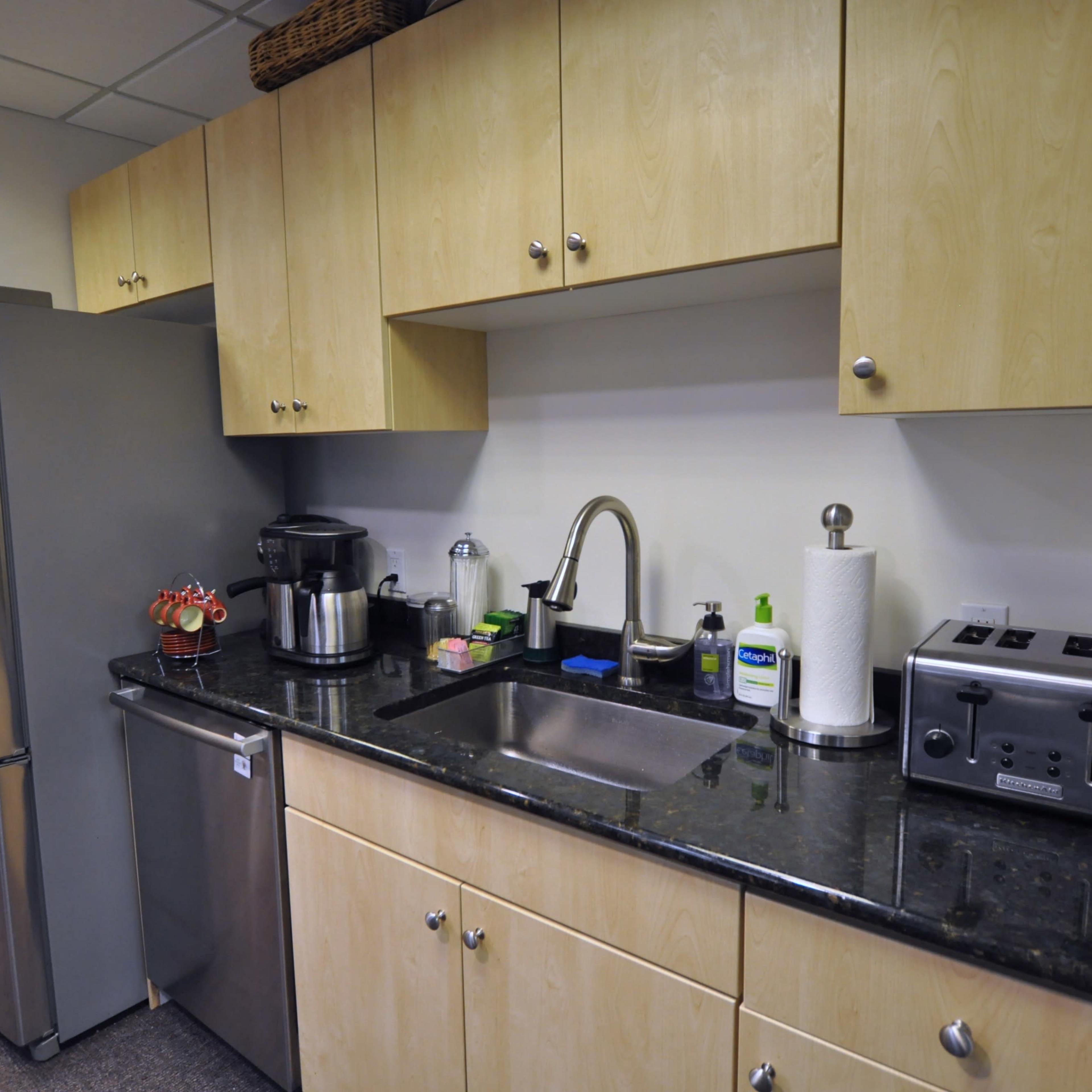 A kitchen area with light wooden cabinets, a stainless steel refrigerator, and a black countertop featuring a sink and various kitchen appliances.