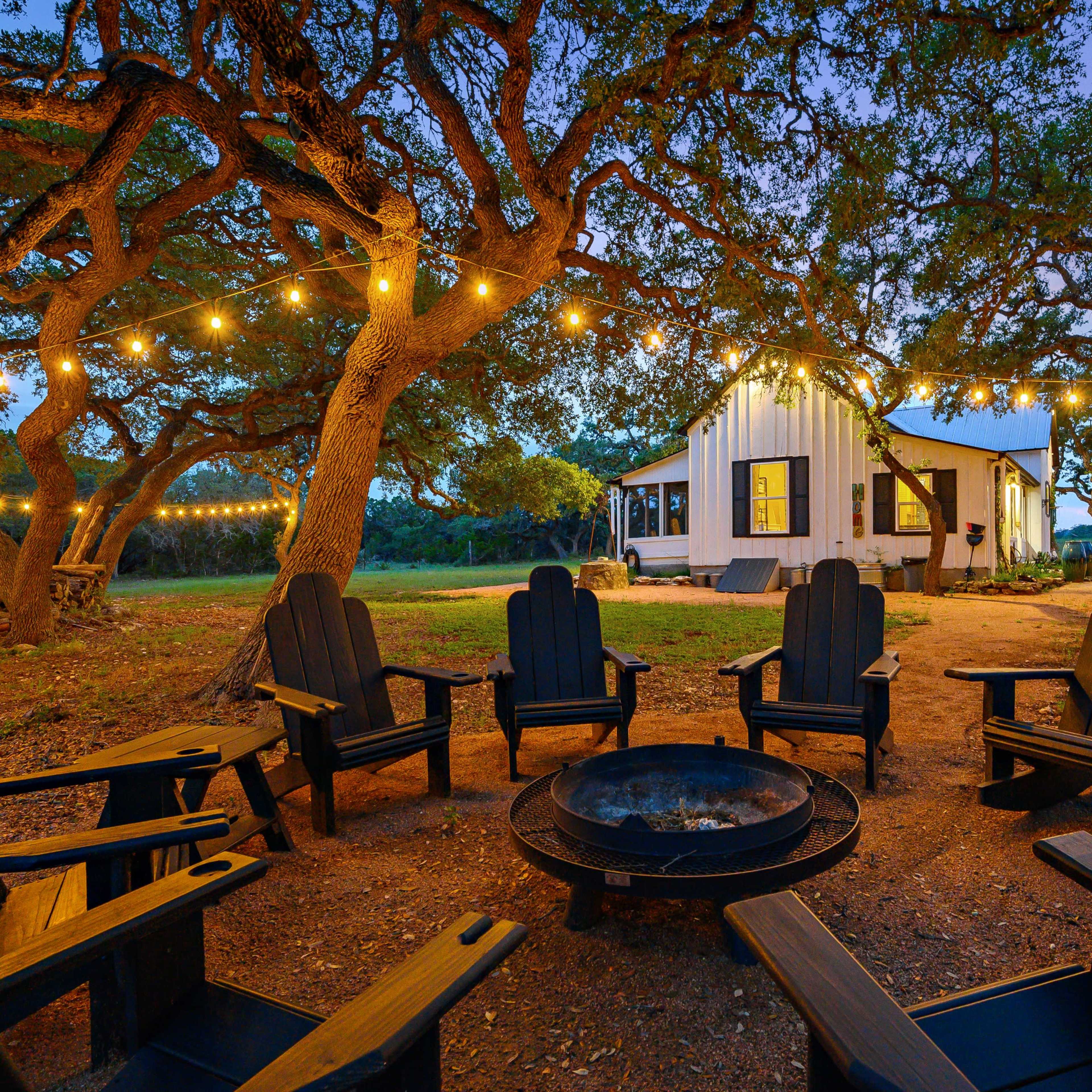 A circle of black Adirondack chairs surrounds a fire pit, with string lights illuminating a white building and trees in the background at dusk.