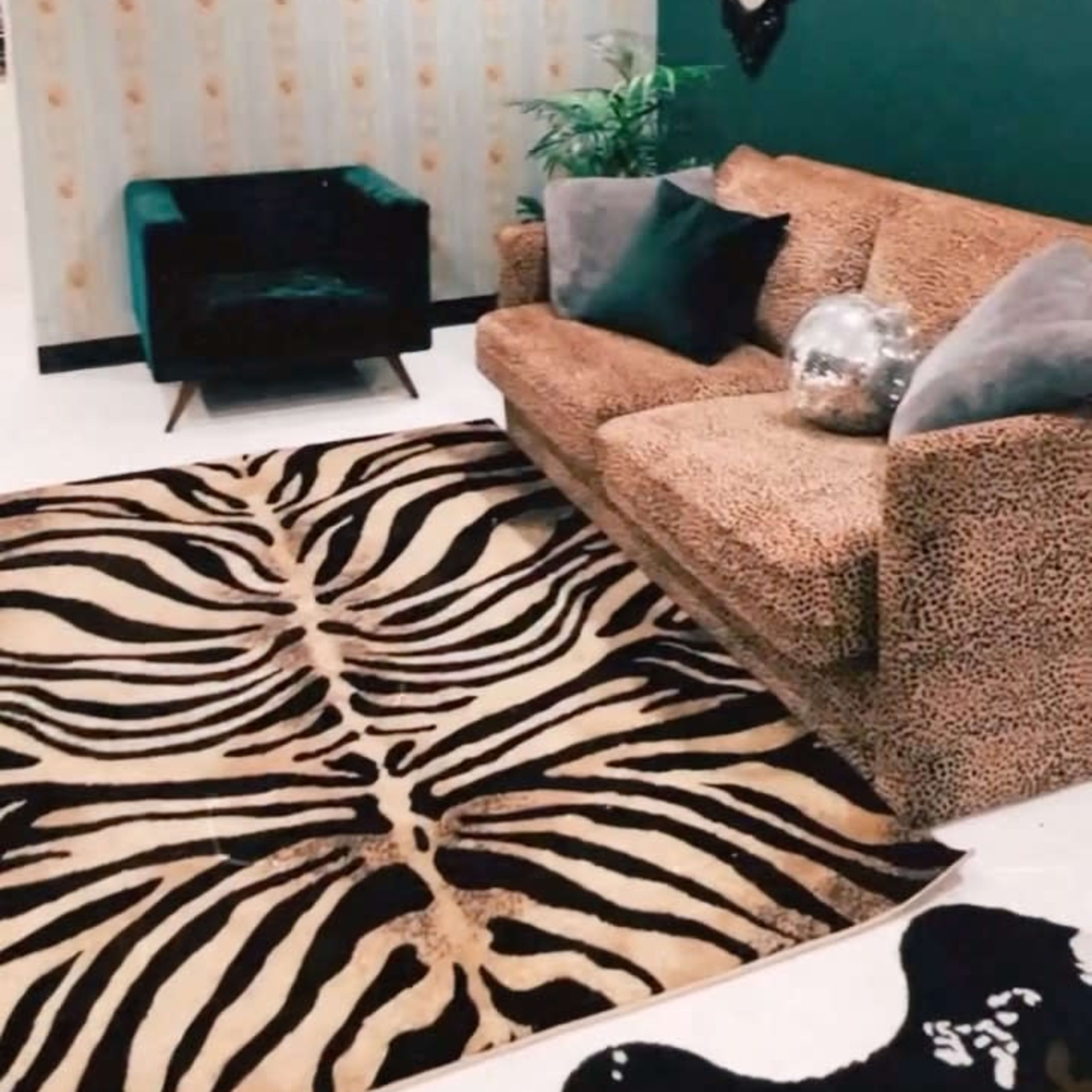 A cozy living room with a leopard print sofa, a black armchair, a zebra-patterned rug, and a wall featuring a decorative mirror and neon artwork.