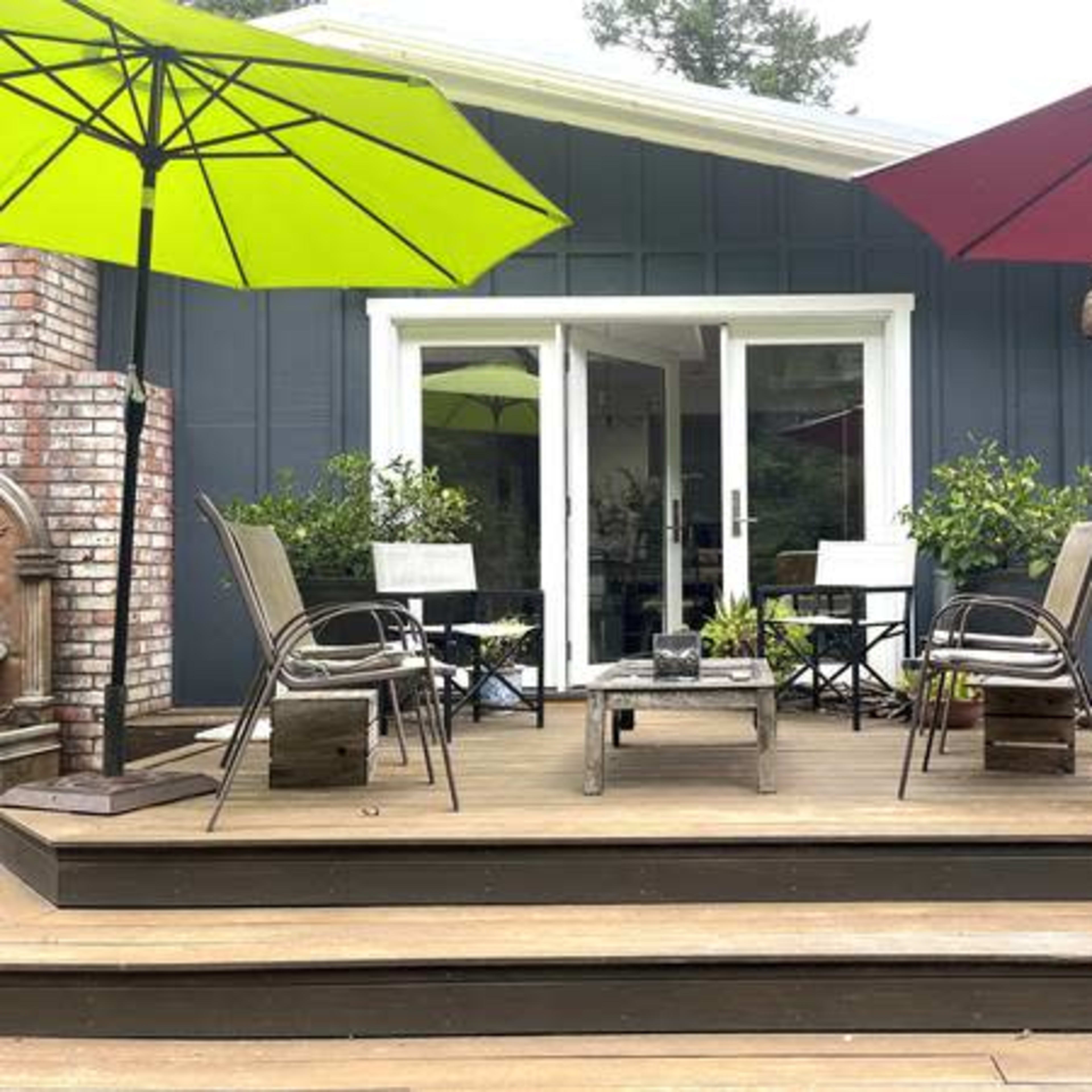A wooden deck with two chairs and a table, surrounded by colorful umbrellas, leading to a house with large windows.