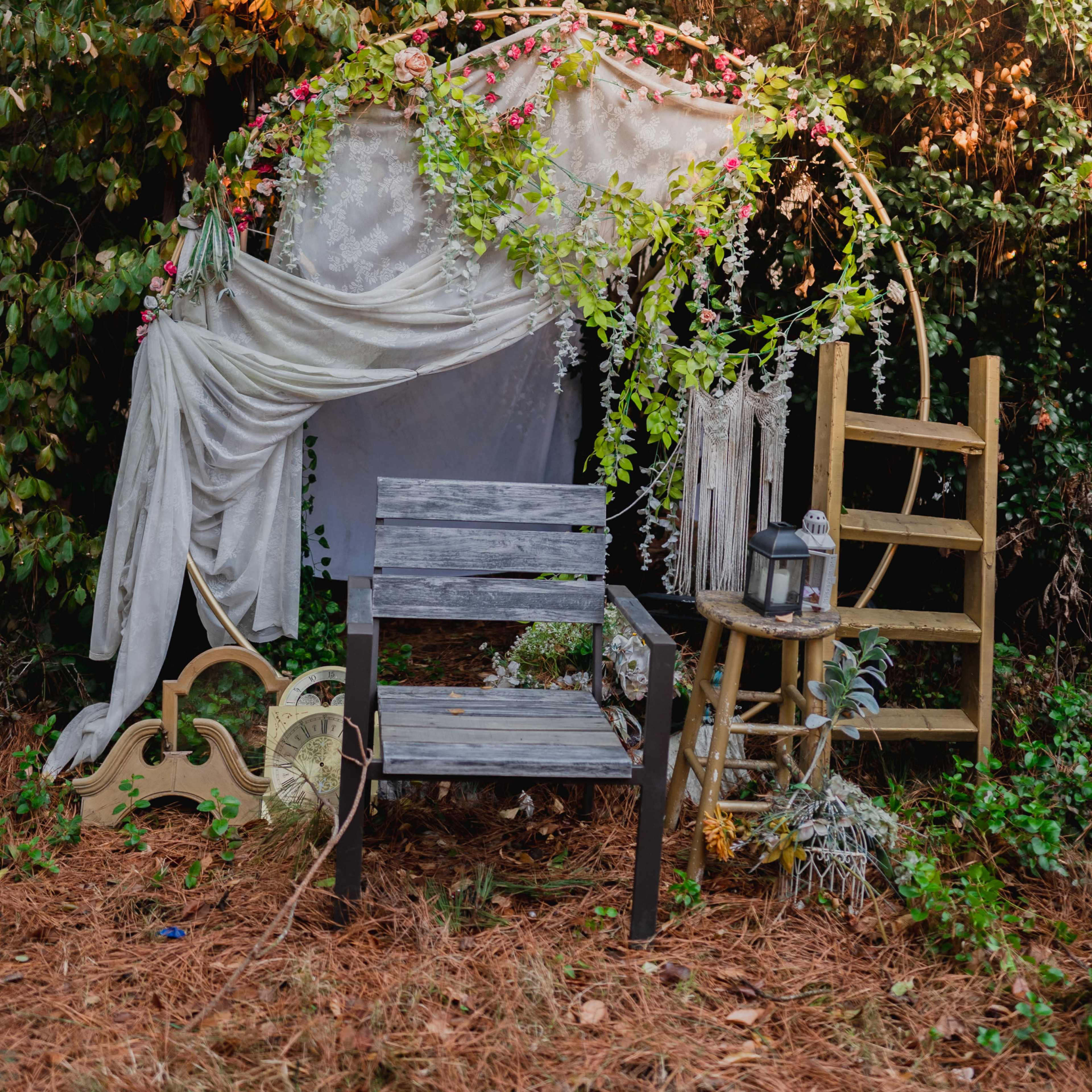 A rustic seating area adorned with floral decorations, a draped fabric canopy, and a wooden ladder, set against a backdrop of greenery.