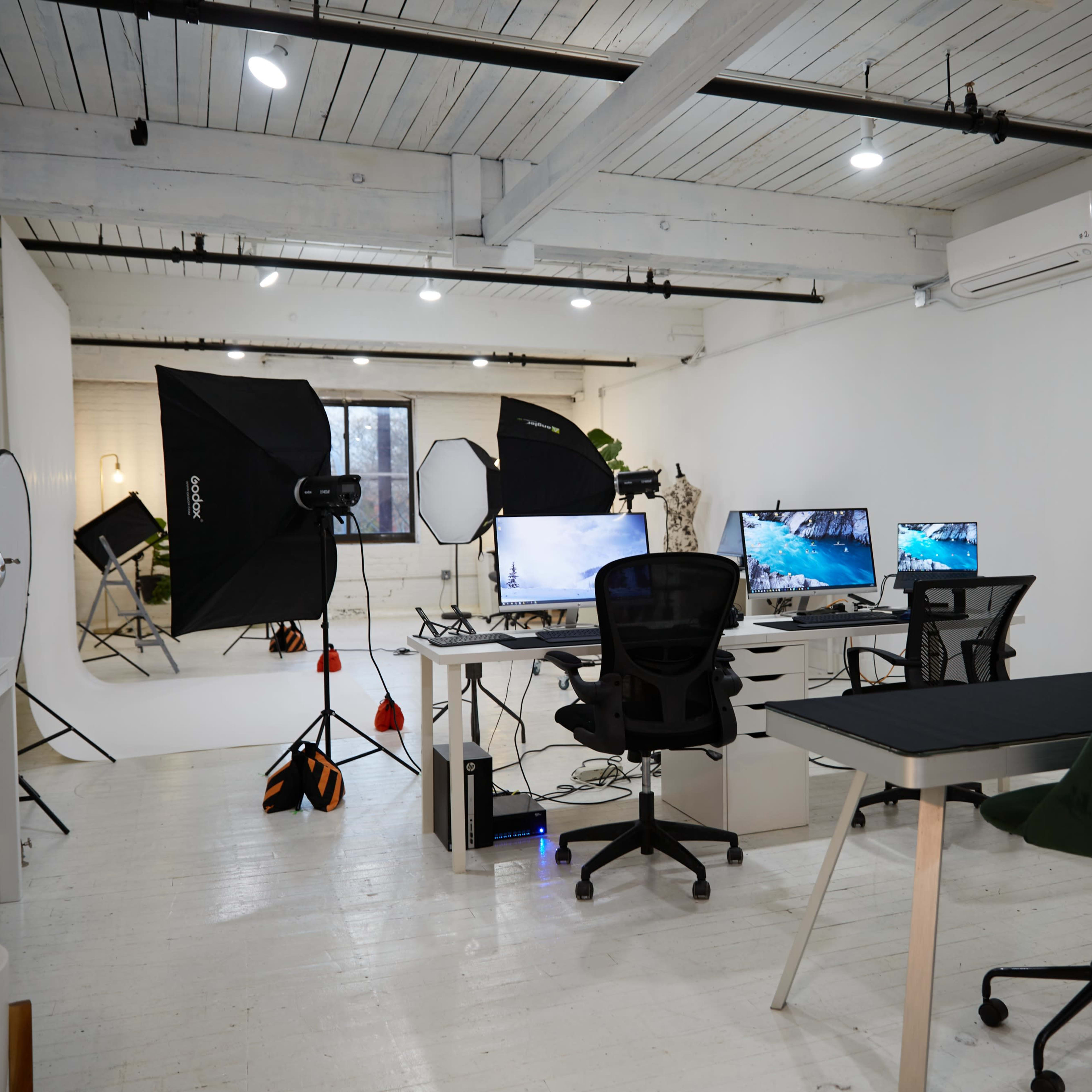 A modern workspace with multiple monitors, office chairs, and studio lighting equipment in a well-lit room with white walls and wooden beams.