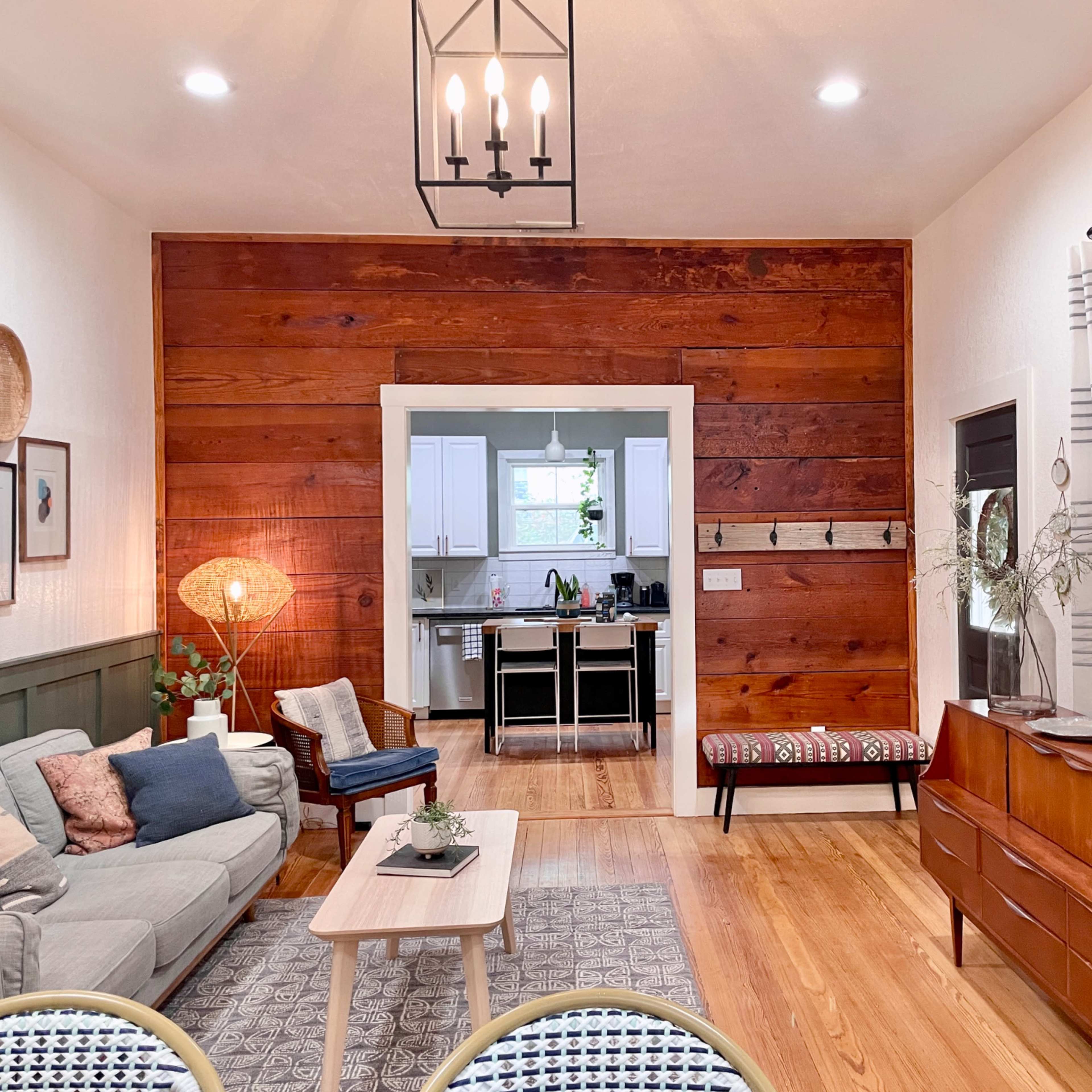 A cozy living room features a gray sofa, wooden accents, and an open view into a kitchen area with modern appliances.