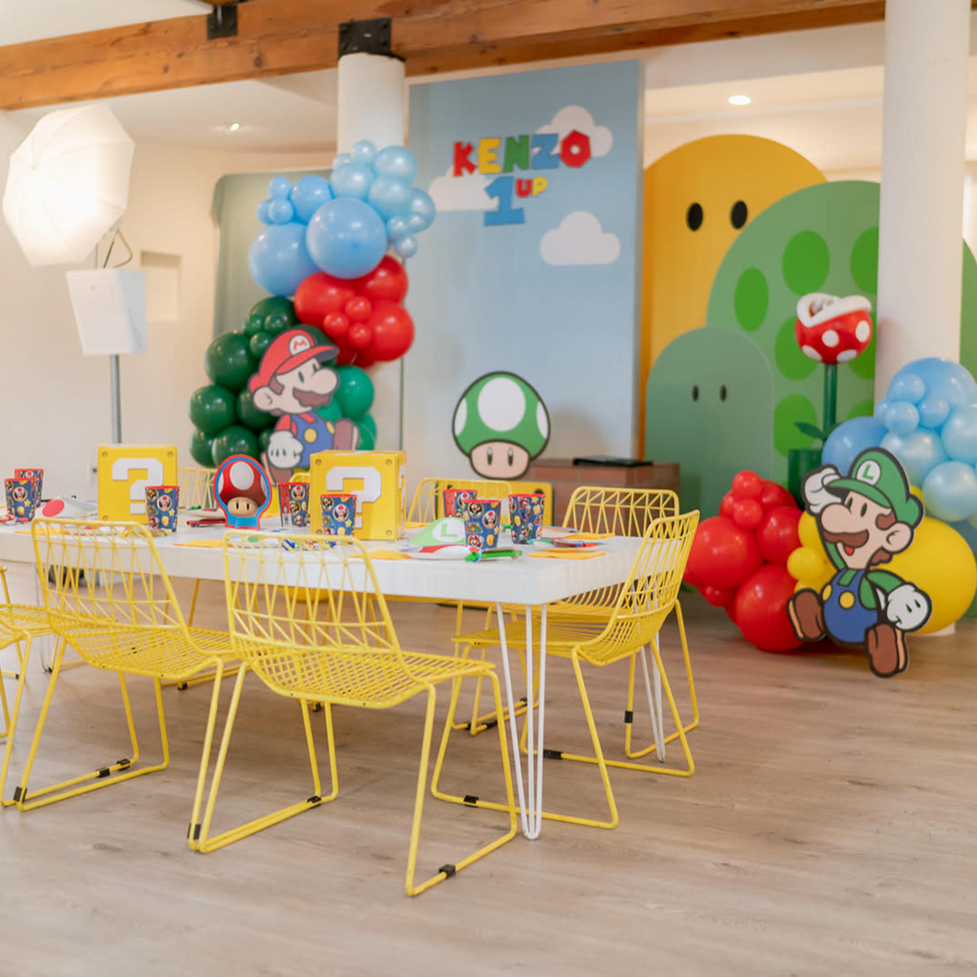 A colorful children's party setup featuring a table surrounded by yellow wire chairs, decorated with balloons and character cutouts from a video game theme.