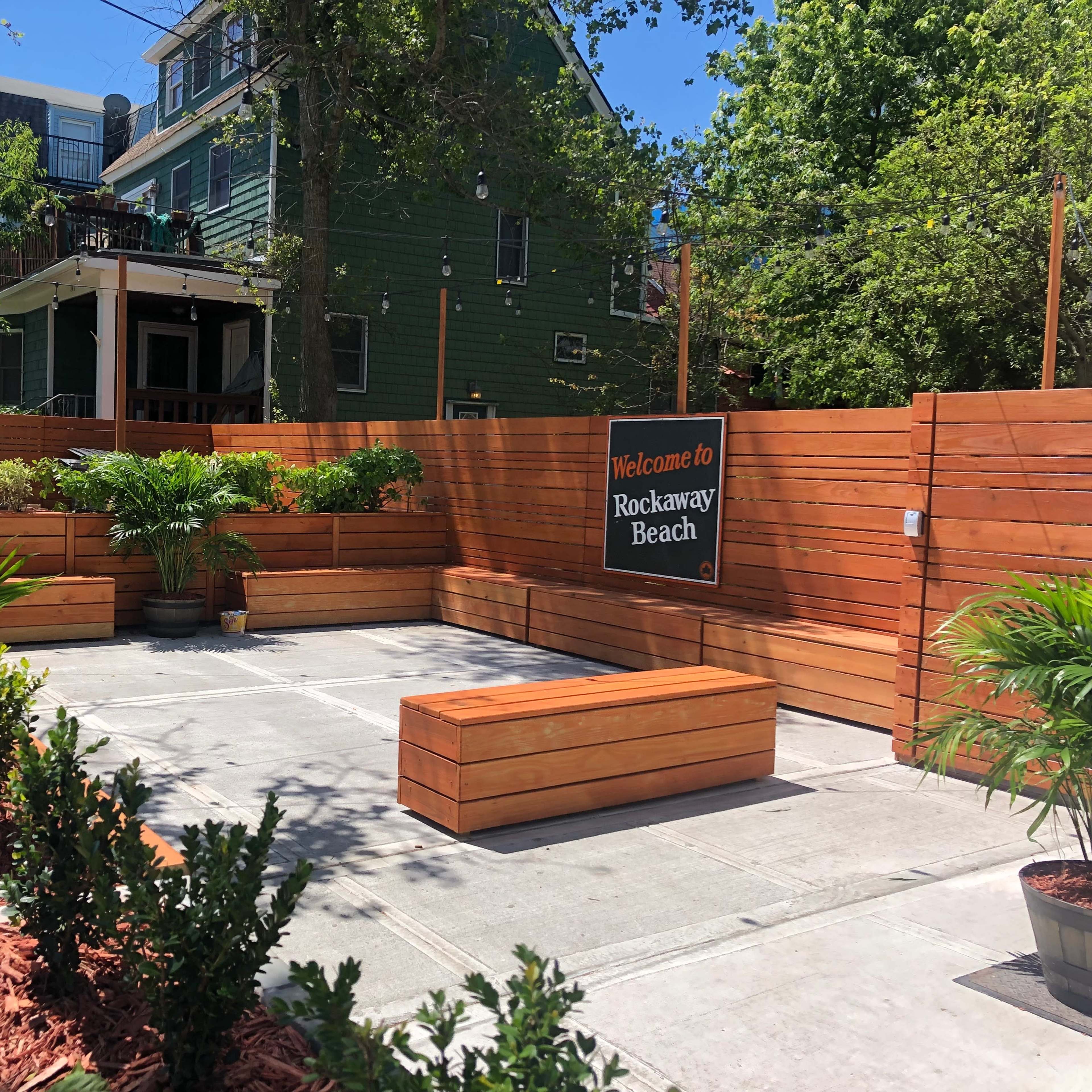 A landscaped outdoor space with wooden benches, potted plants, and a sign that reads "Welcome to Rockaway Beach."