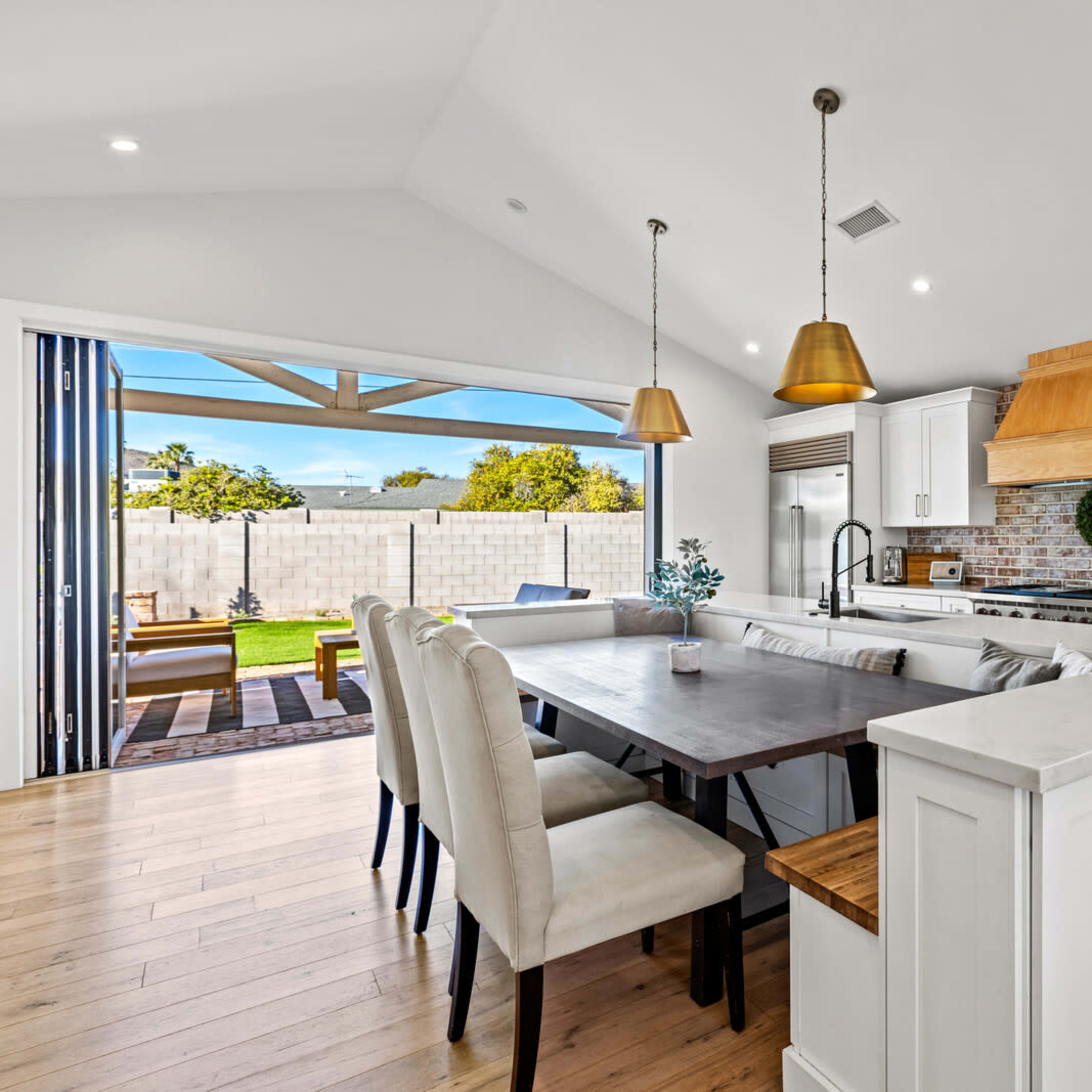 A modern kitchen features a large dining table, white cabinetry, wooden accents, and pendant lights, with a view of a backyard through sliding glass doors.