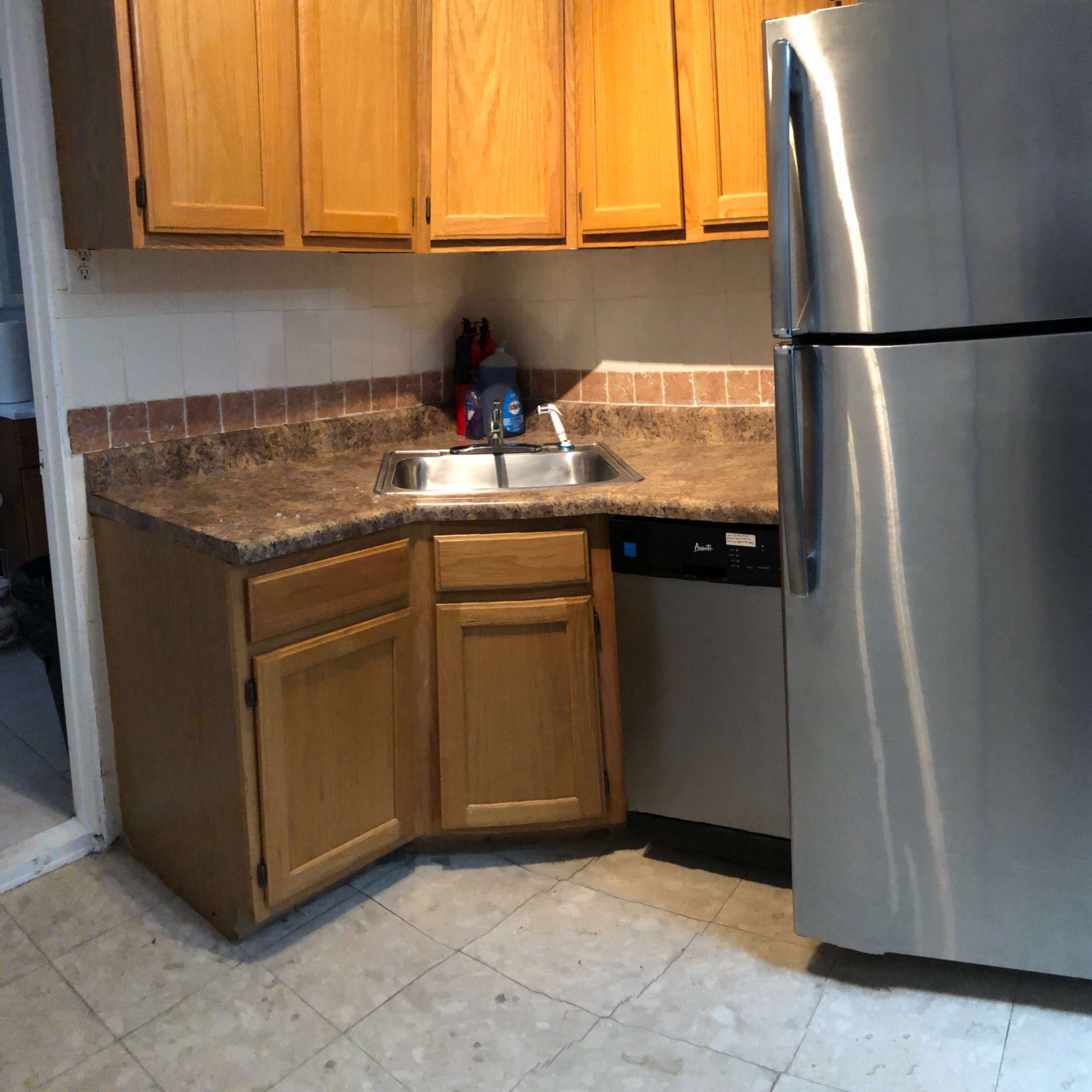 A kitchen corner with wooden cabinets, a stainless steel refrigerator, a sink, and a dishwasher.