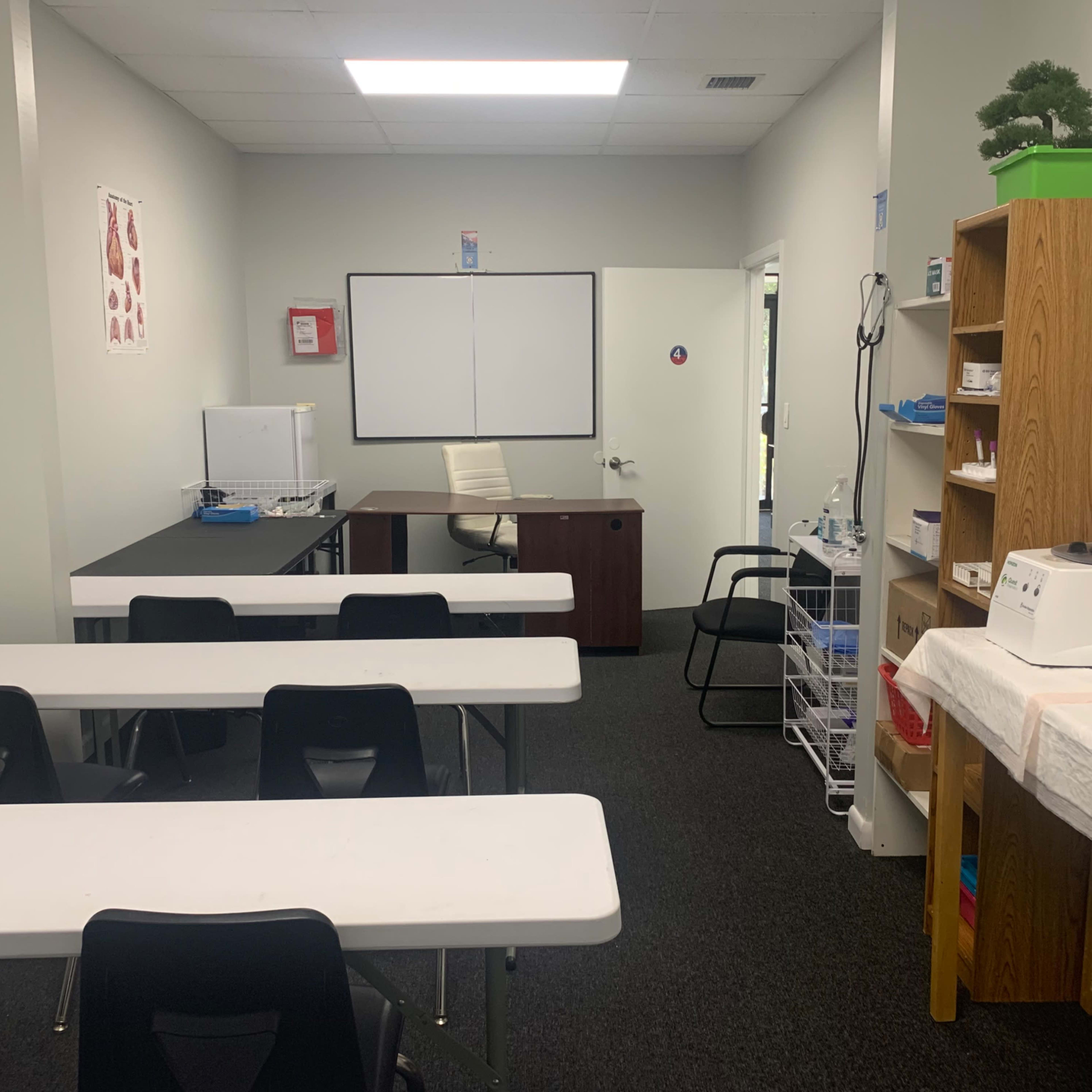 A classroom with several white tables and black chairs, a desk in the corner, and shelves containing various items.