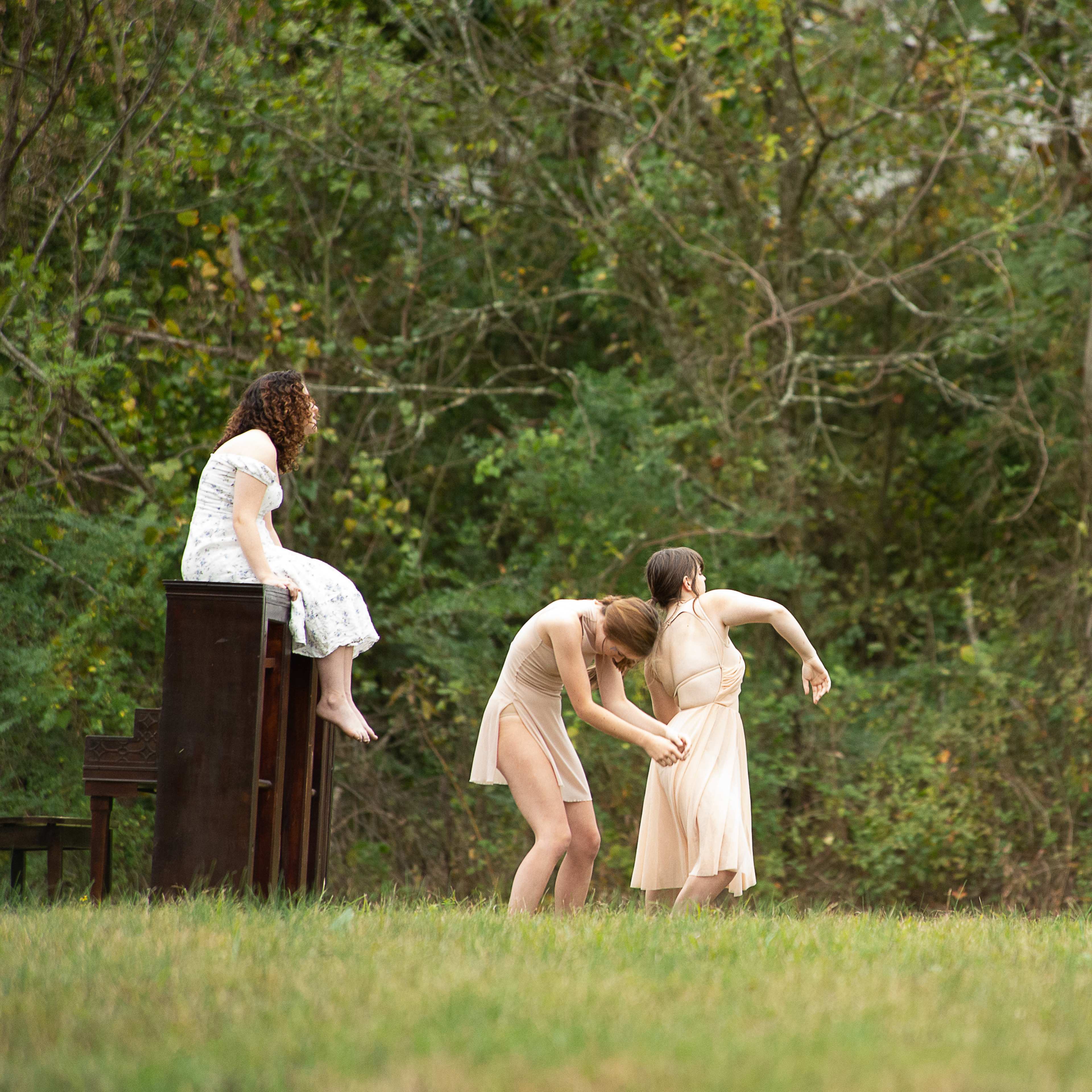 A woman sits on an upright piano while two dancers perform nearby, and a man records the scene with a camera in a natural outdoor setting.