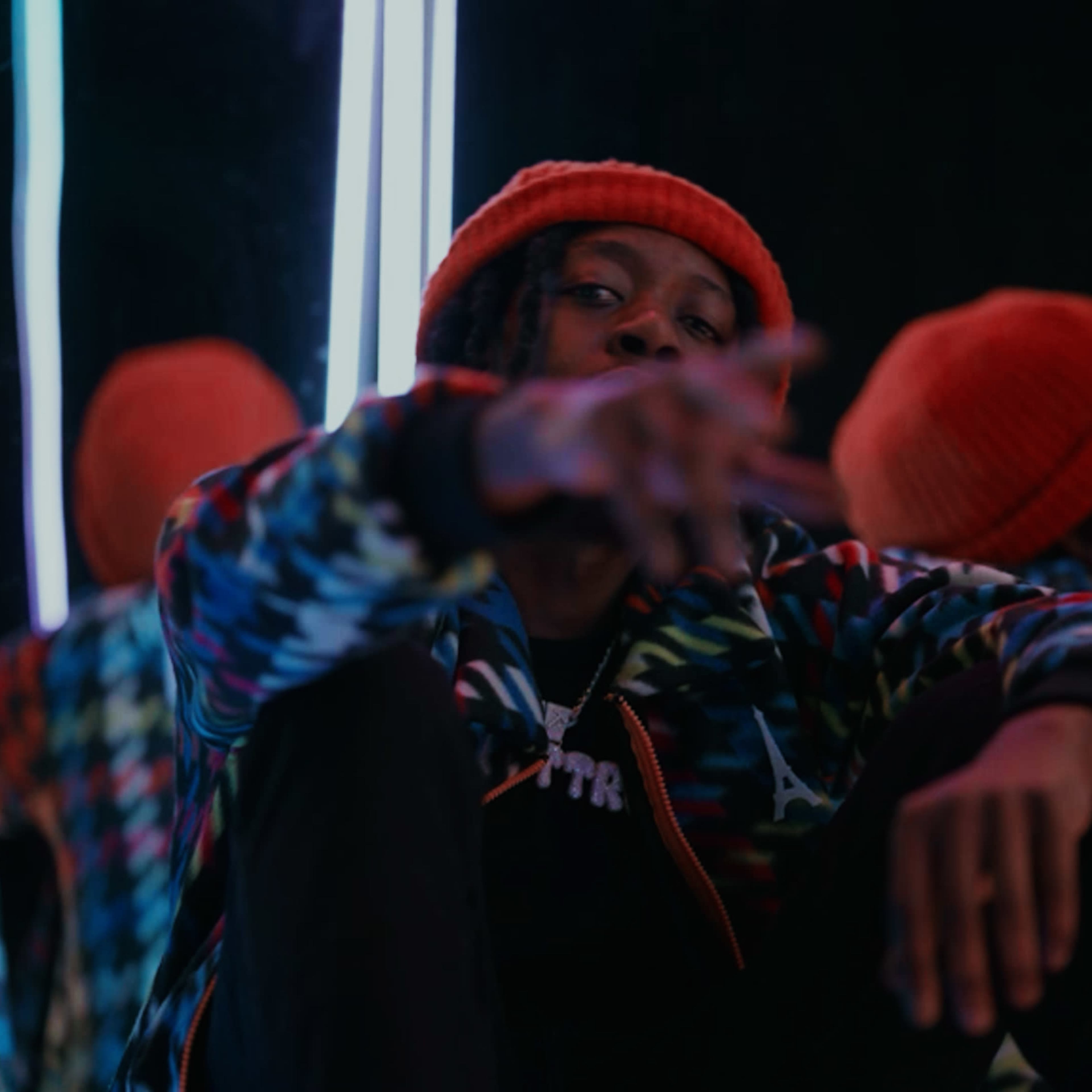 A person in a colorful jacket and orange beanie poses in front of mirrors with bright neon lights.