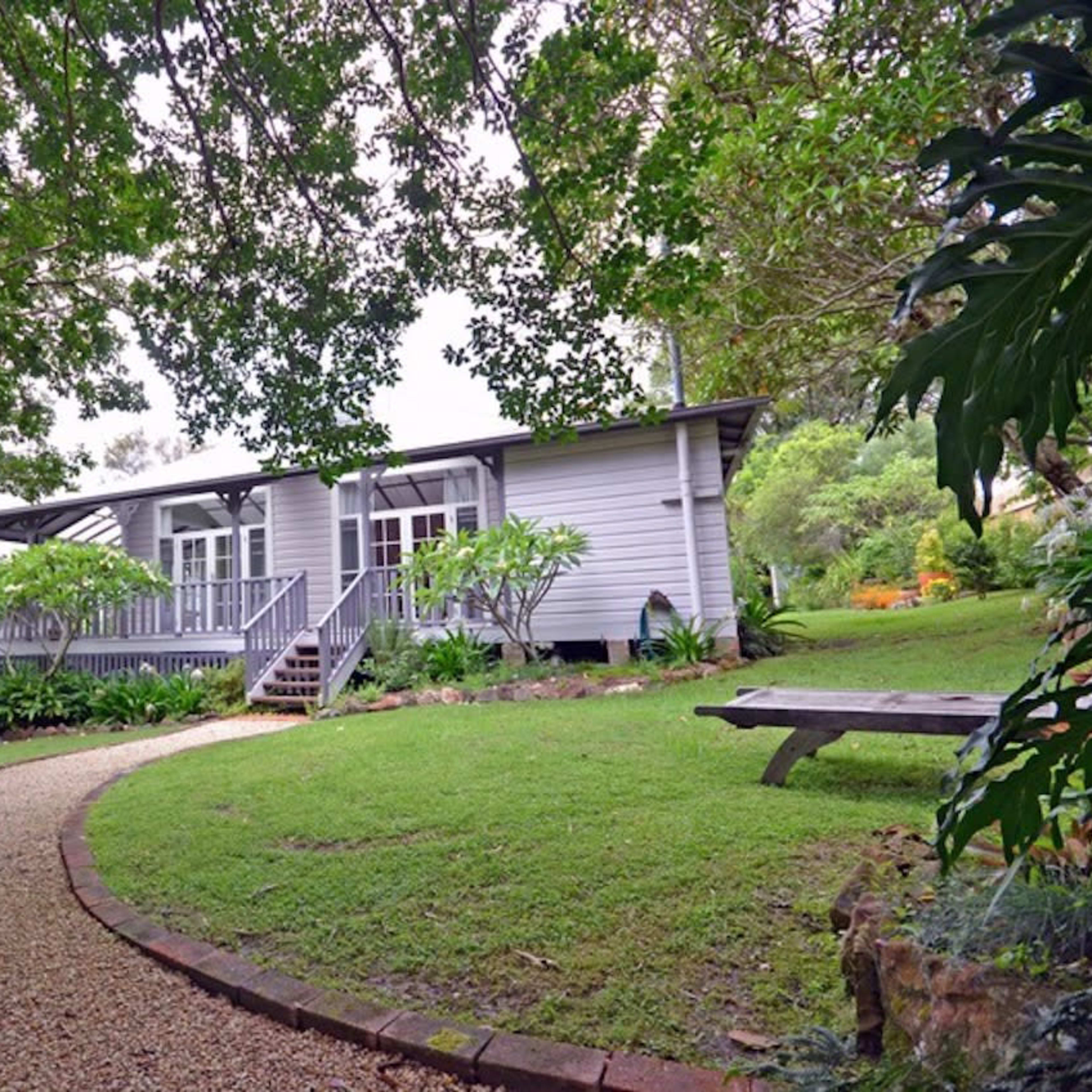A single-story house with large windows is surrounded by lush greenery and a gravel pathway leading to the entrance.