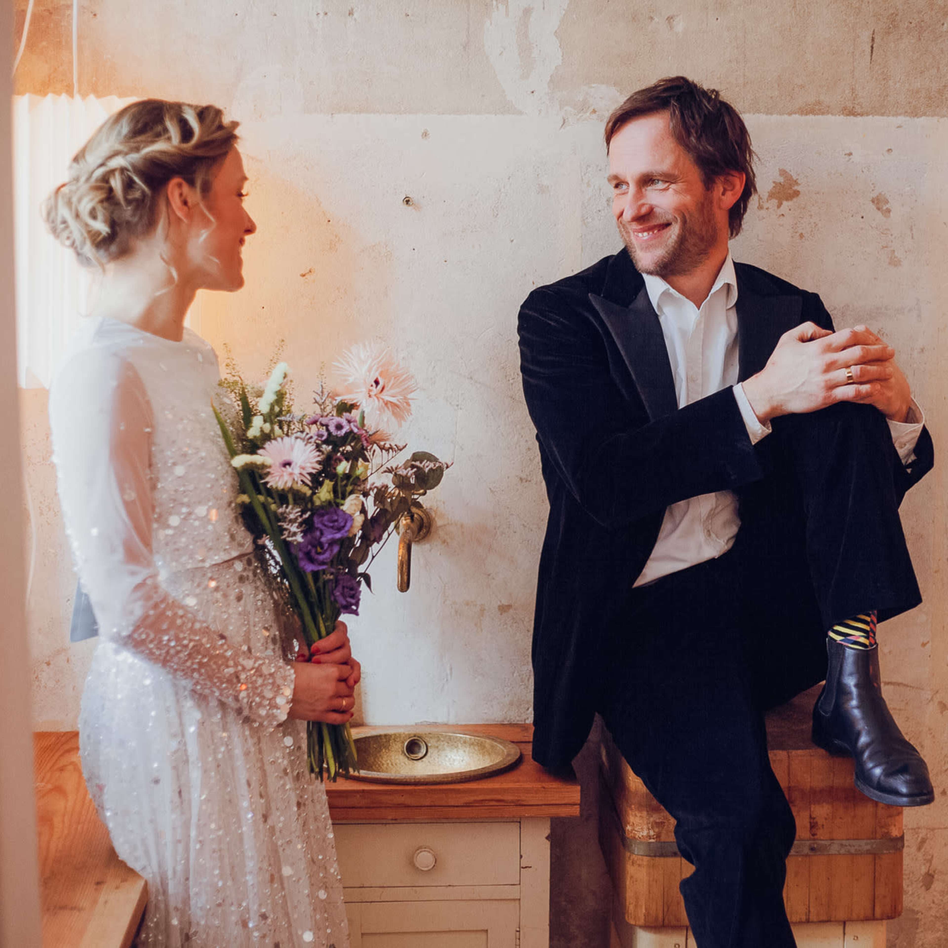 A woman in a sequined dress stands holding a bouquet of flowers while a man in a black jacket sits on a wooden crate, both smiling at each other in a rustic room.