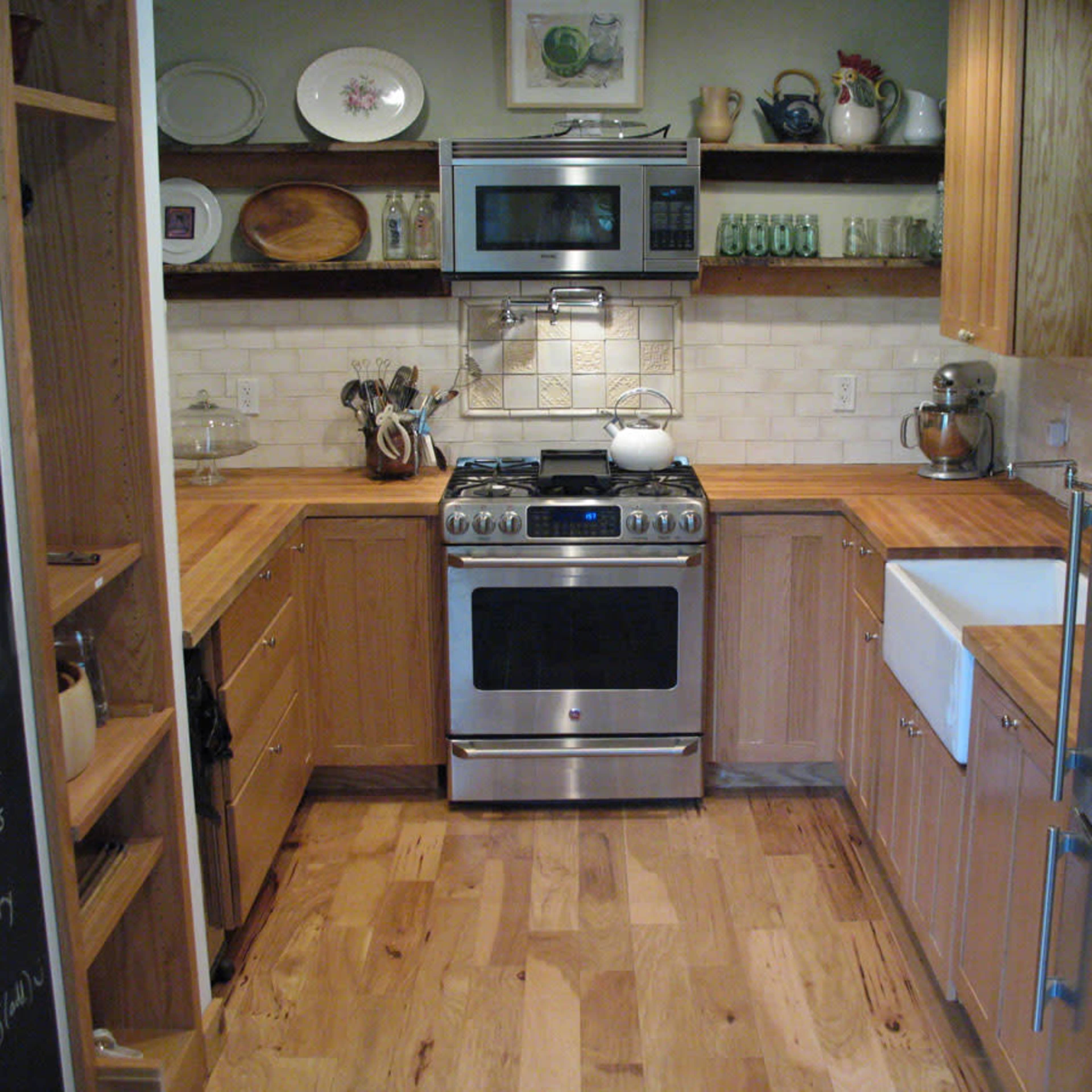 A compact kitchen featuring wooden cabinetry, a stainless steel oven, and a large farmhouse sink, with open shelving displaying dishes and glassware.