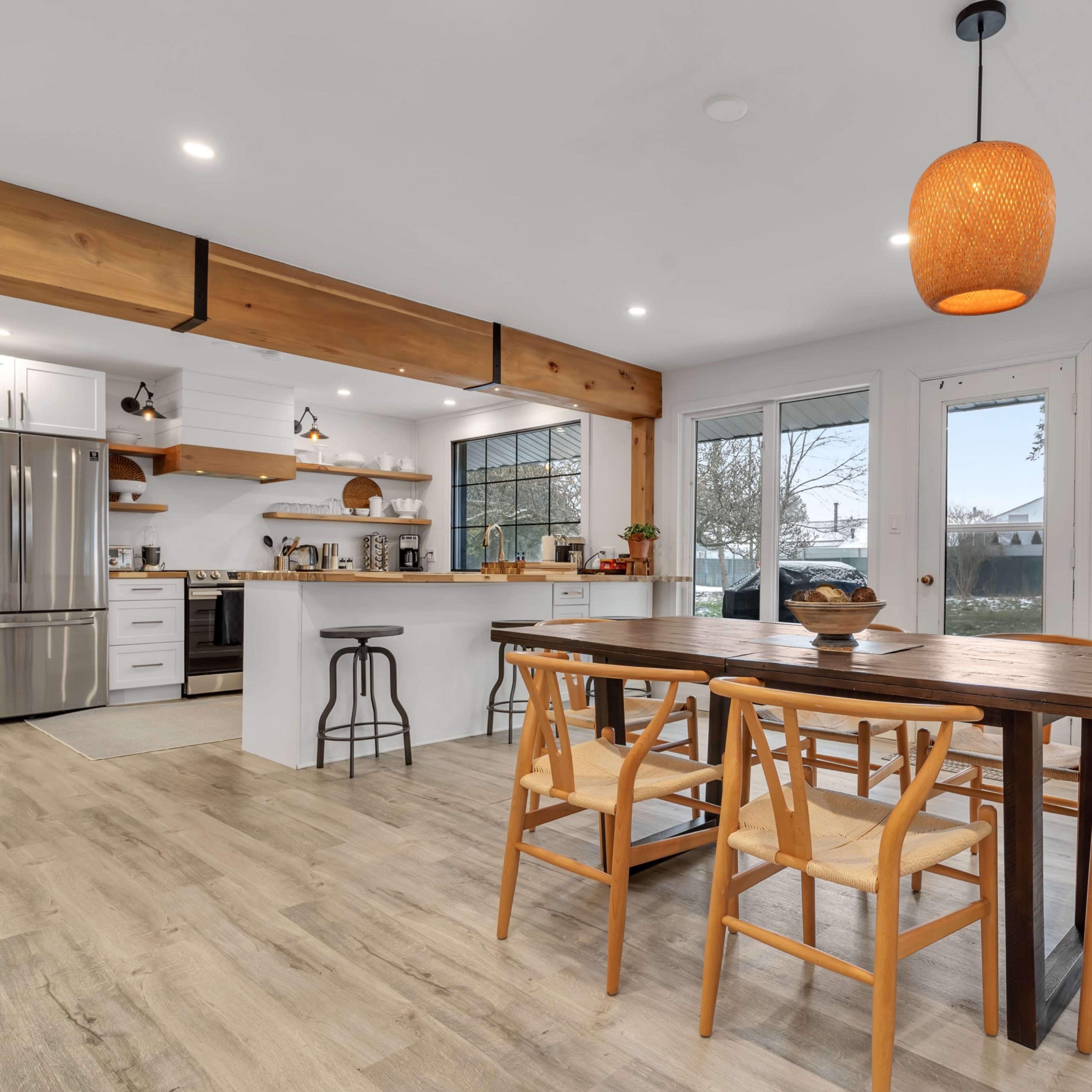A modern kitchen features a wooden dining table surrounded by chairs, with a large window illuminating stainless steel appliances and open shelving.