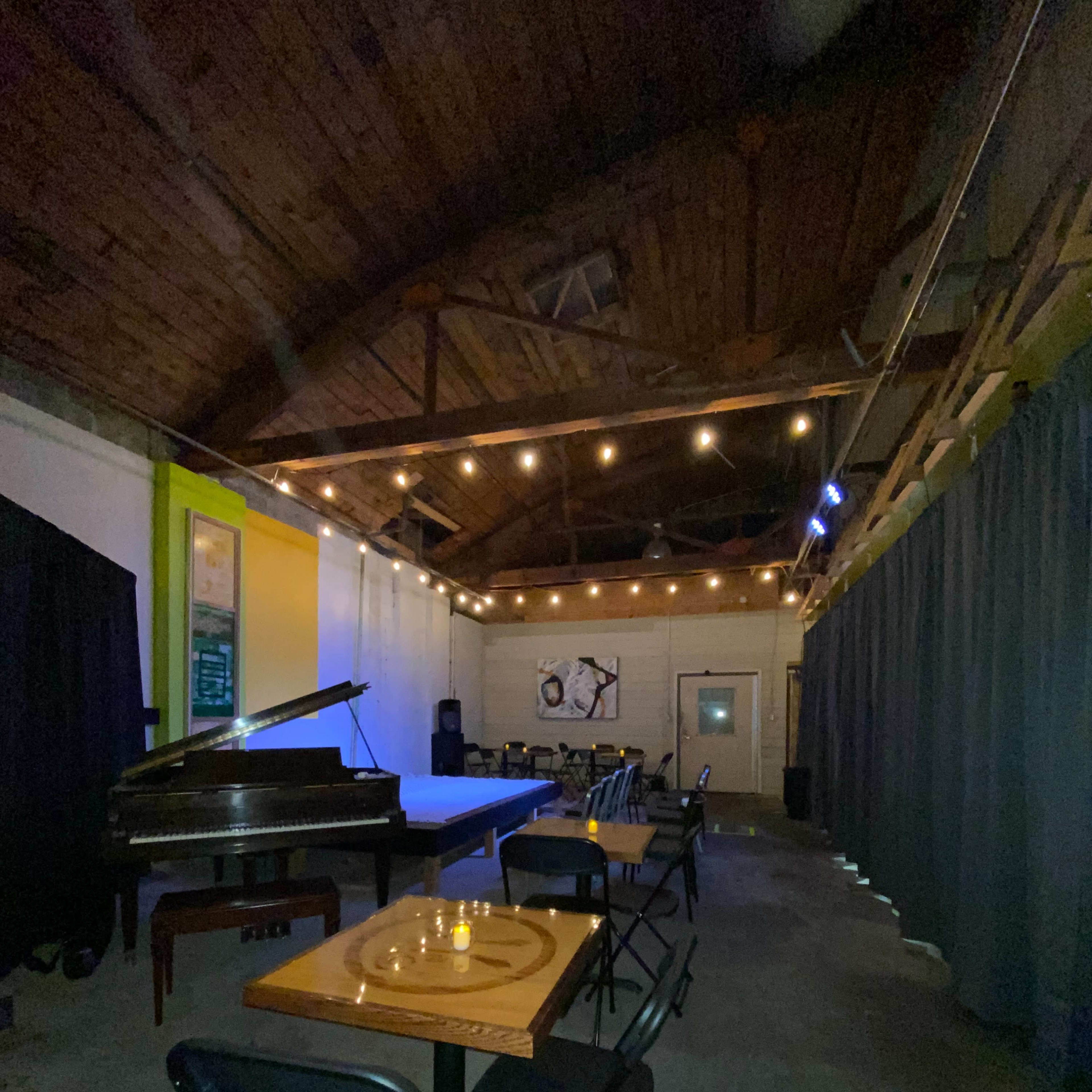 An empty performance space with a grand piano, tables set for guests, and overhead string lights illuminating the wooden ceiling.