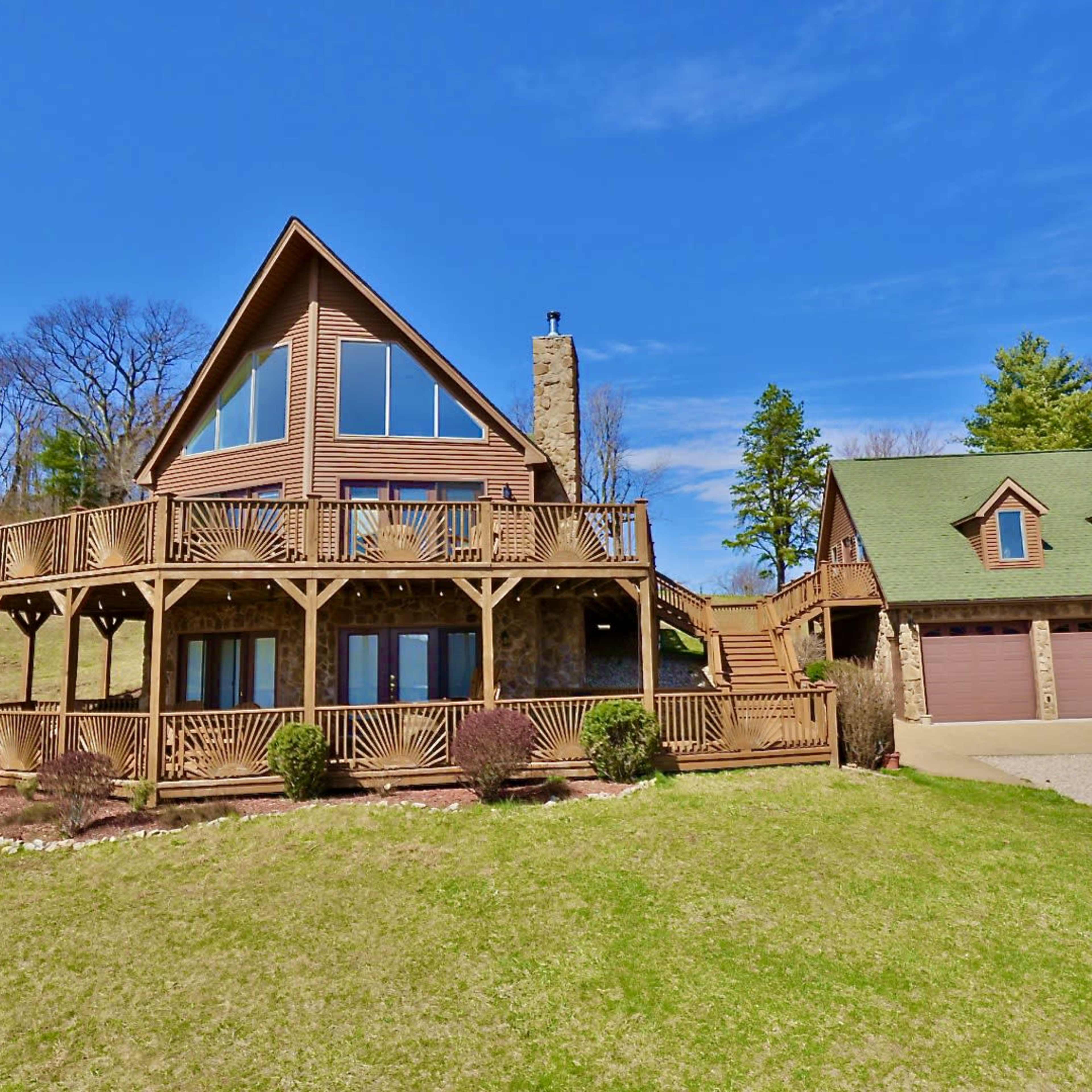 A multi-level house with large windows and a wraparound deck is situated on a grassy hillside, accompanied by a two-car garage.