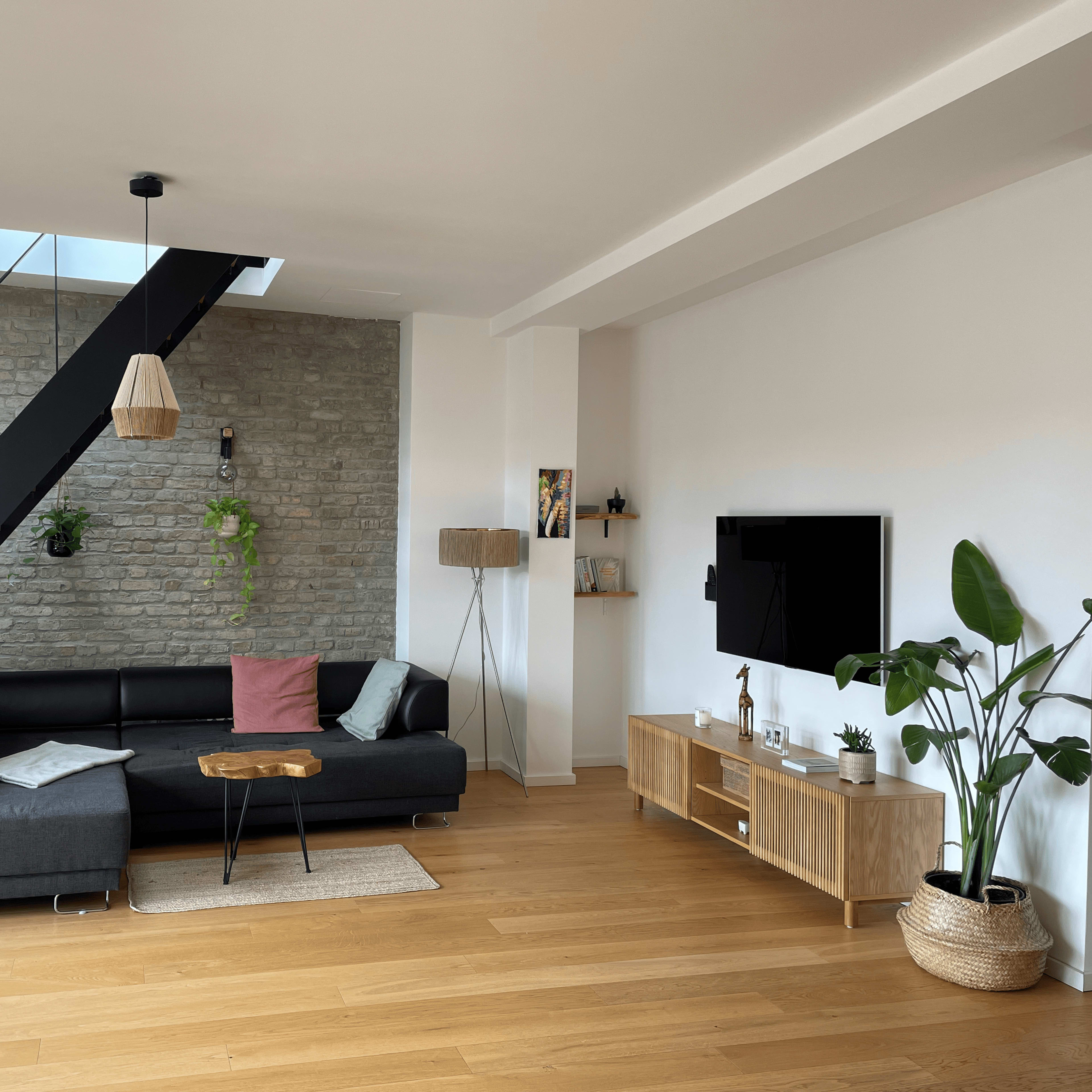 A modern living room featuring a black sectional sofa, a wooden coffee table, a television mounted on a wall, and plants in decorative pots.