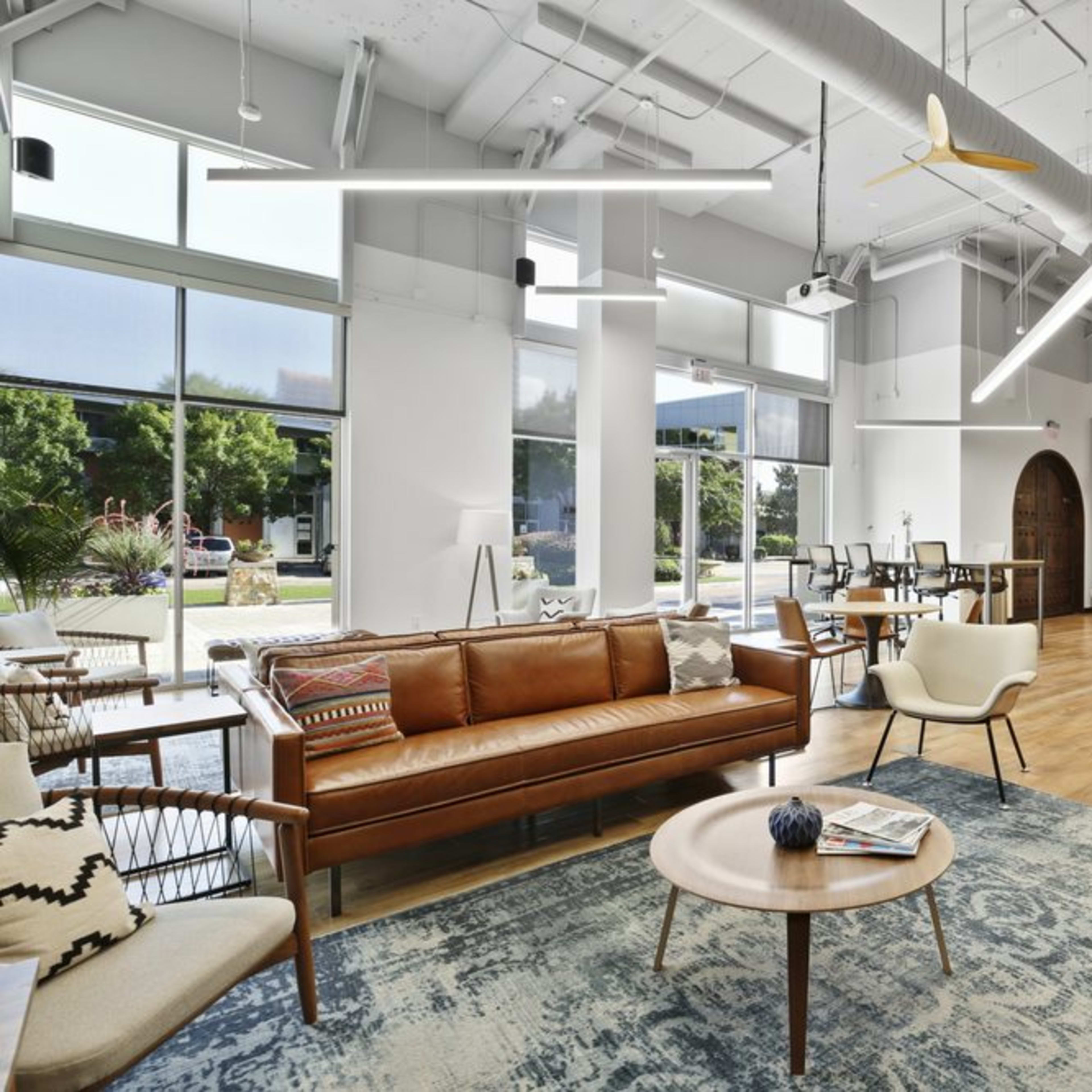 A modern and spacious communal lounge area furnished with a leather sofa, wooden coffee table, and various seating arrangements, alongside large windows that let in natural light.

