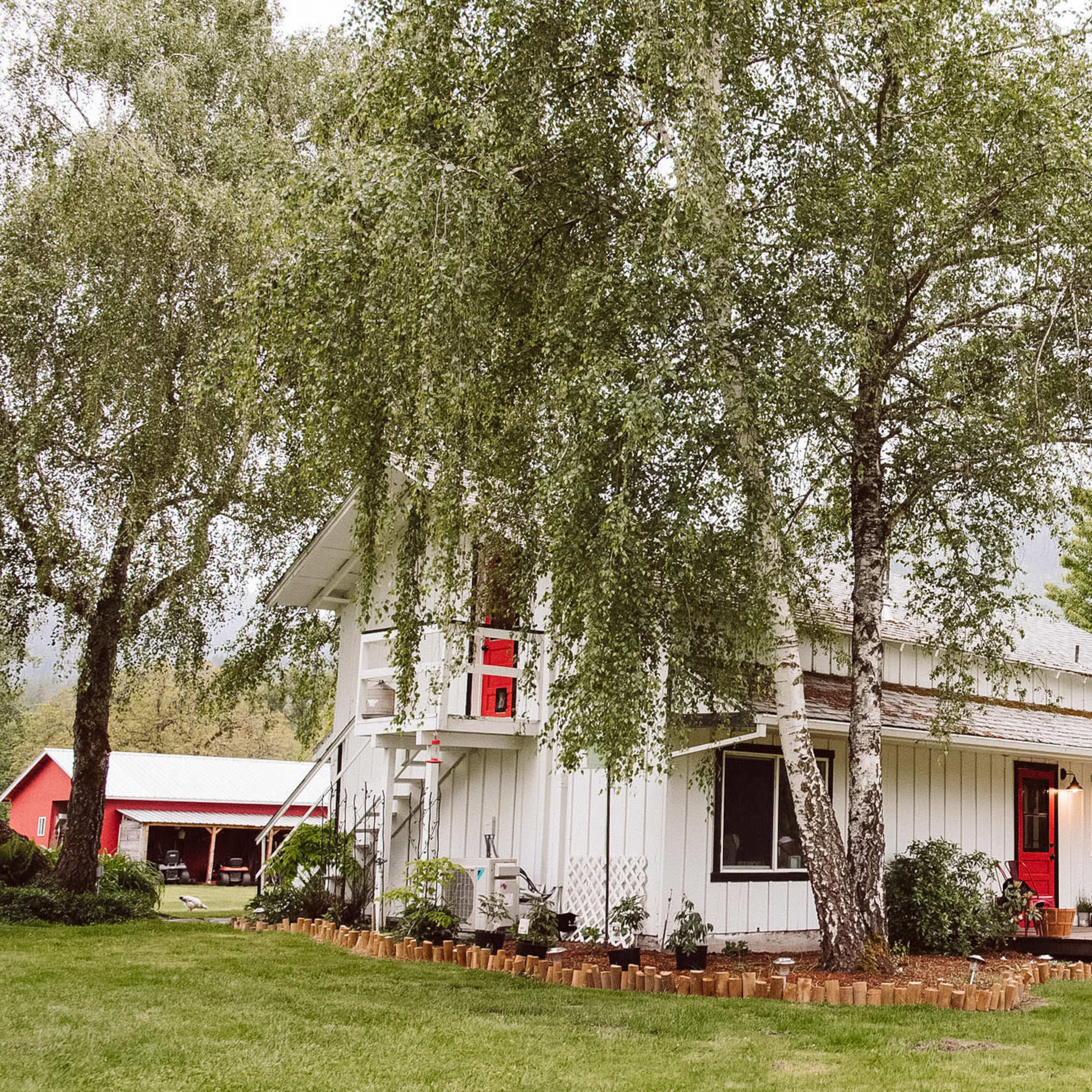 A white two-story house with red accents is surrounded by greenery and a well-maintained lawn.