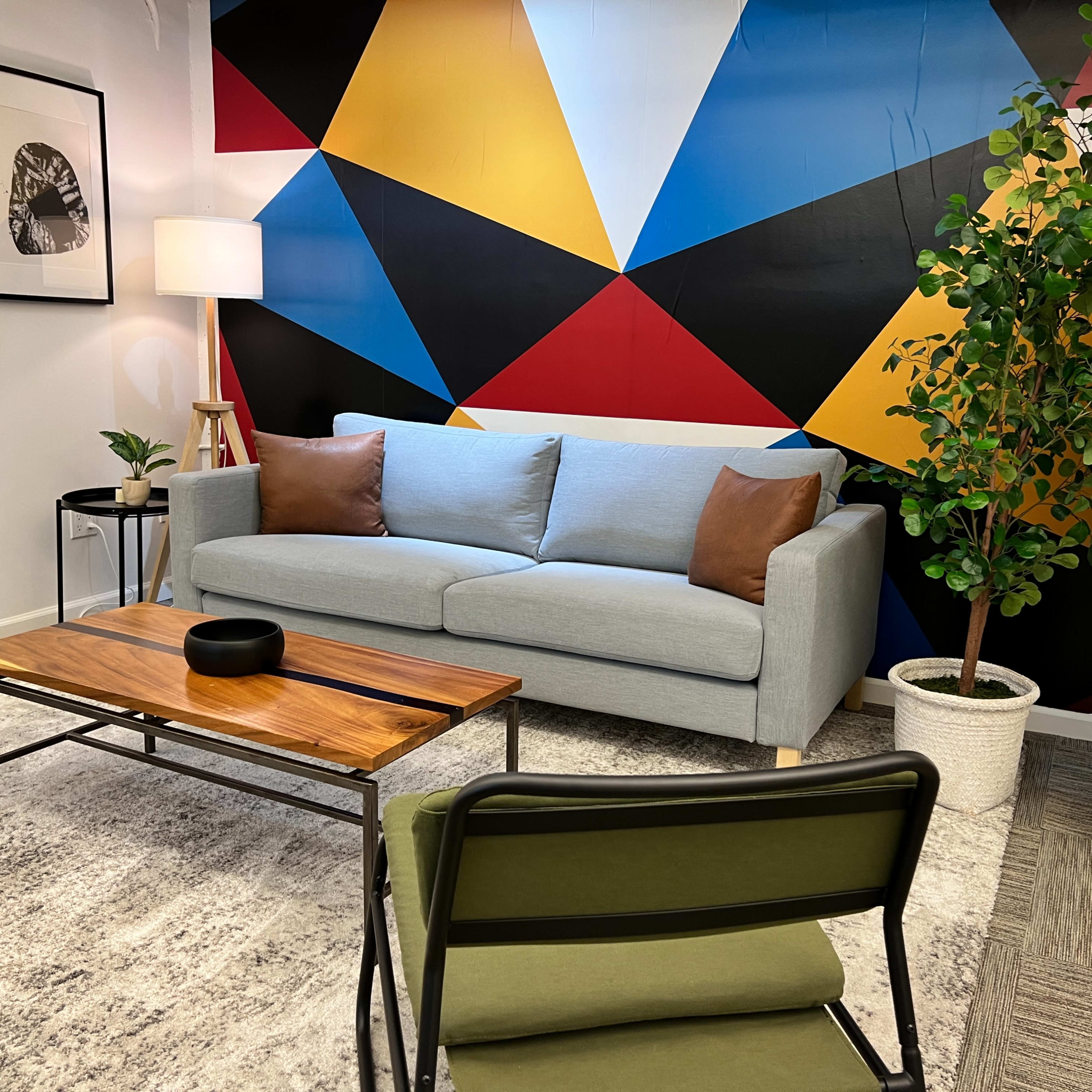 A modern seating area with a light blue sofa, a wooden coffee table, and a patterned mural on the wall, complemented by a lamp and a potted plant.