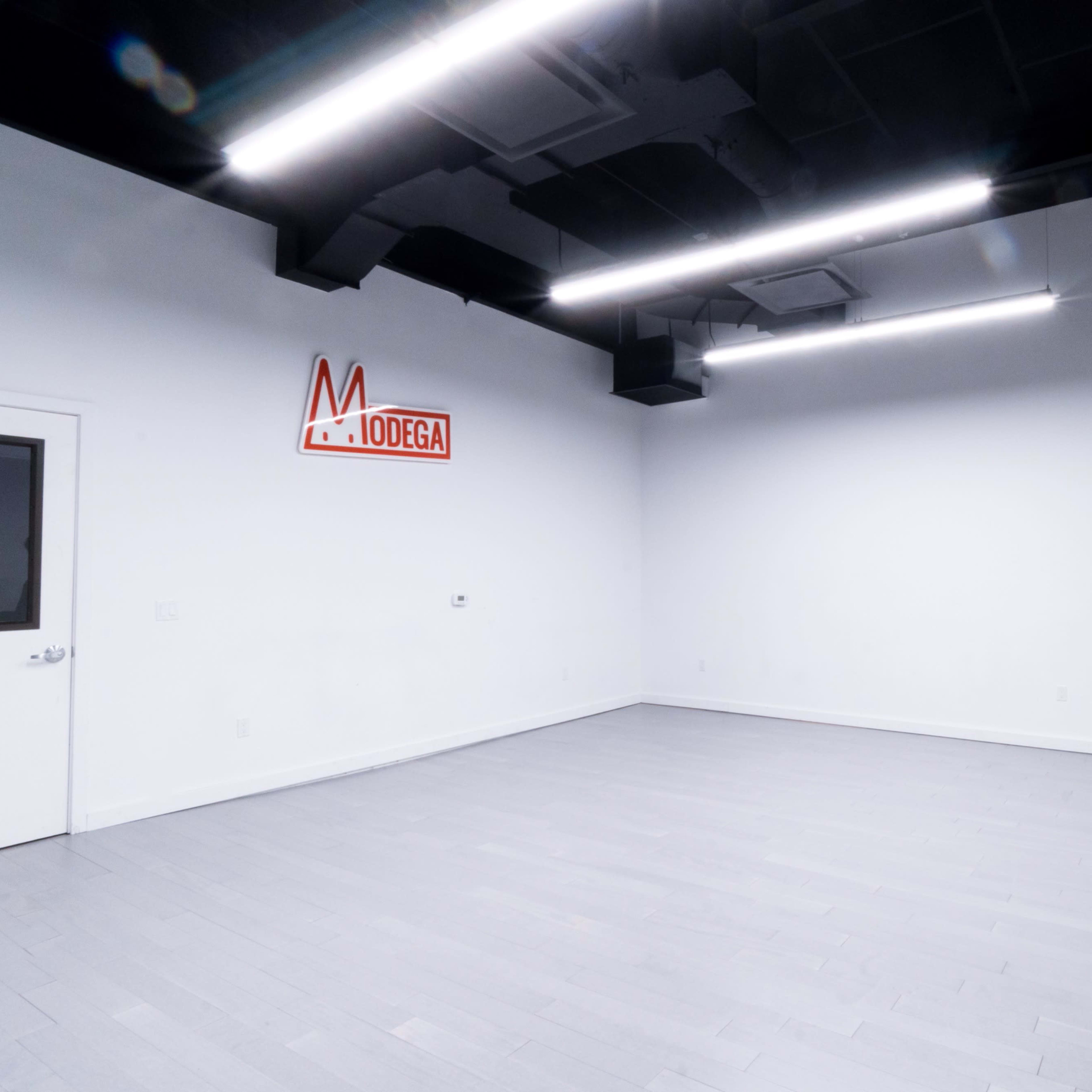 A nearly empty room with white walls, gray flooring, a door on the left, and a large mirror along the right side, featuring a neon sign that reads "MODEGA."