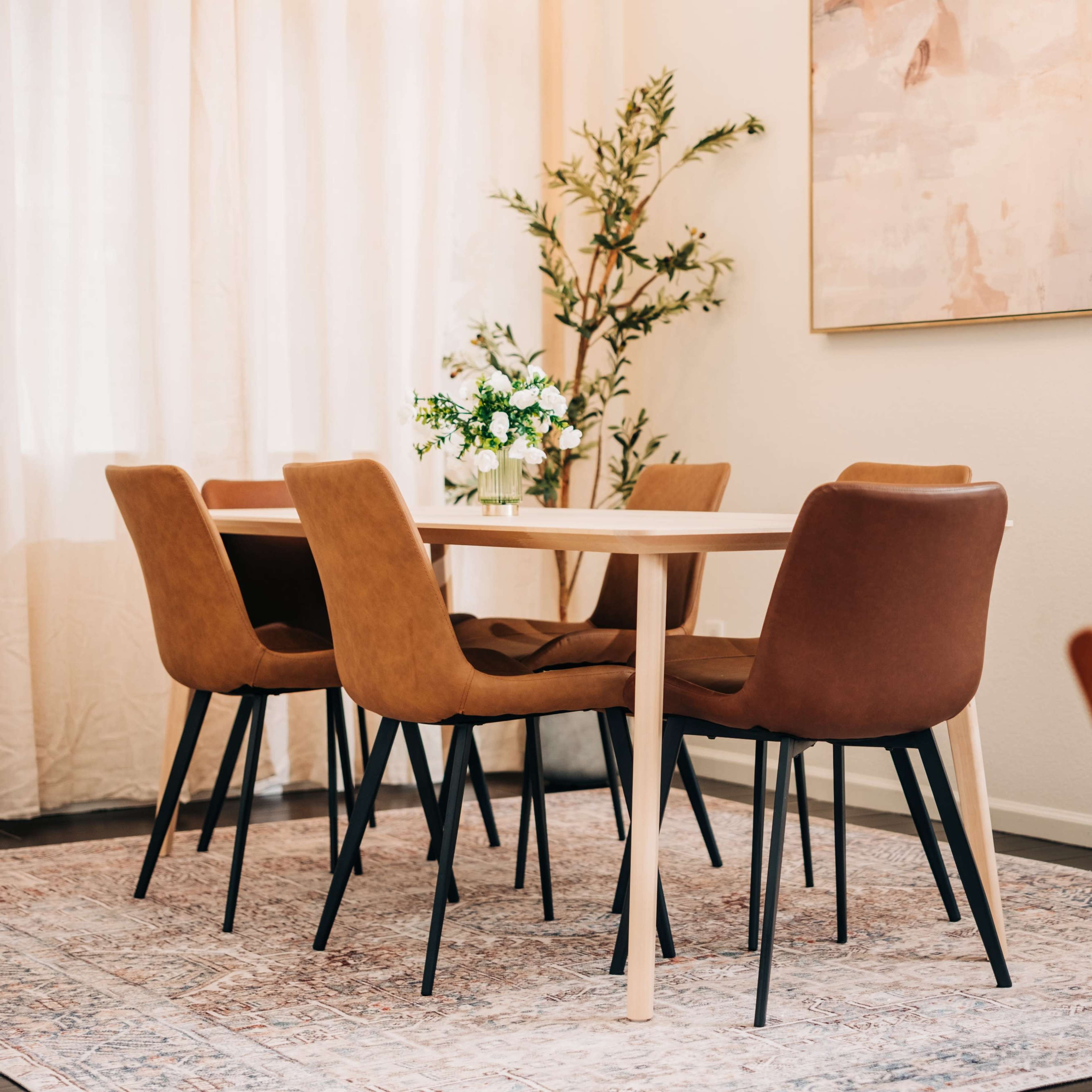 A dining area features a wooden table surrounded by four brown chairs, with a small floral arrangement on the table and a large artwork on the wall.