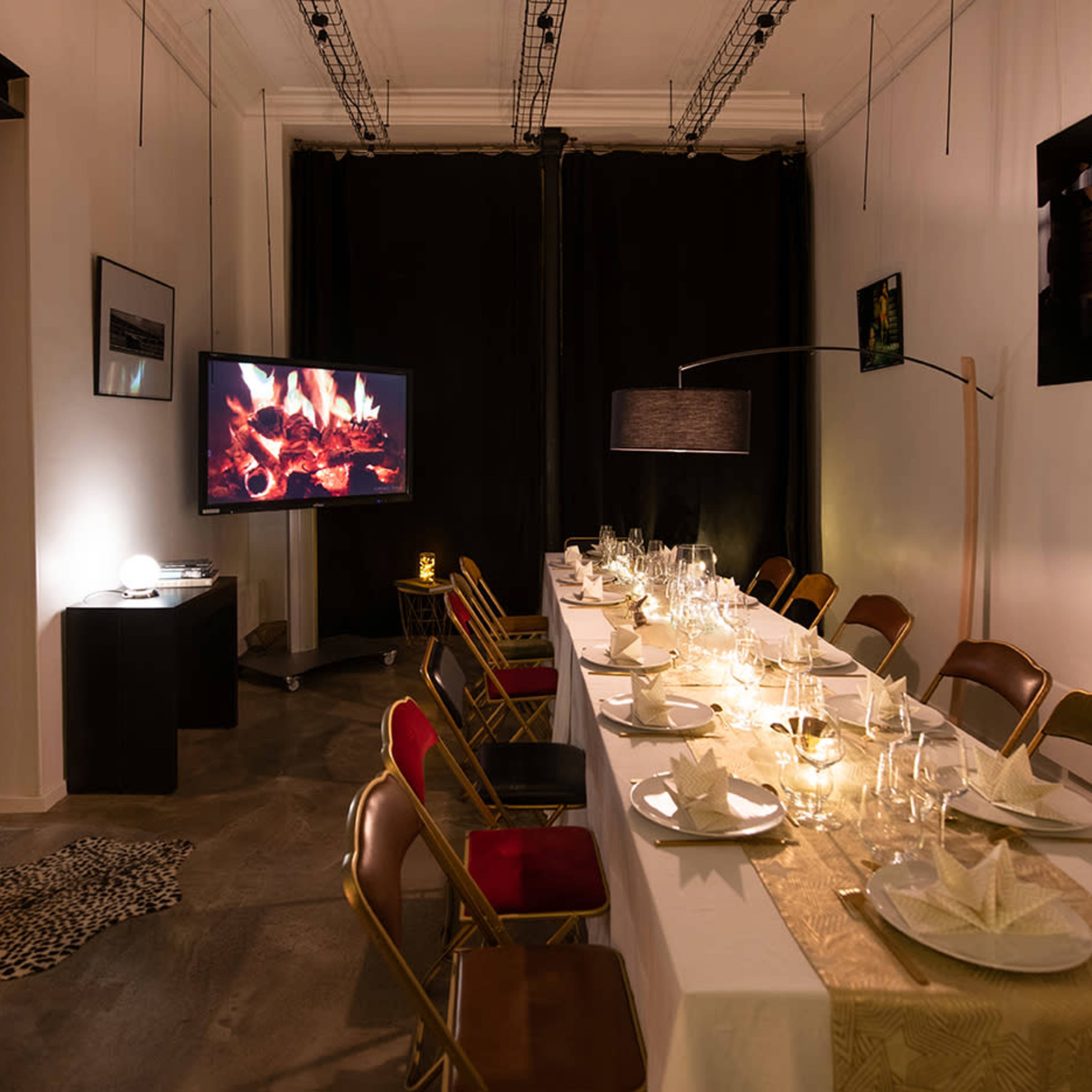 The scene shows a dining area set with a long table adorned with plates and glasses, flanked by chairs, and featuring a television displaying a fireplace backdrop.