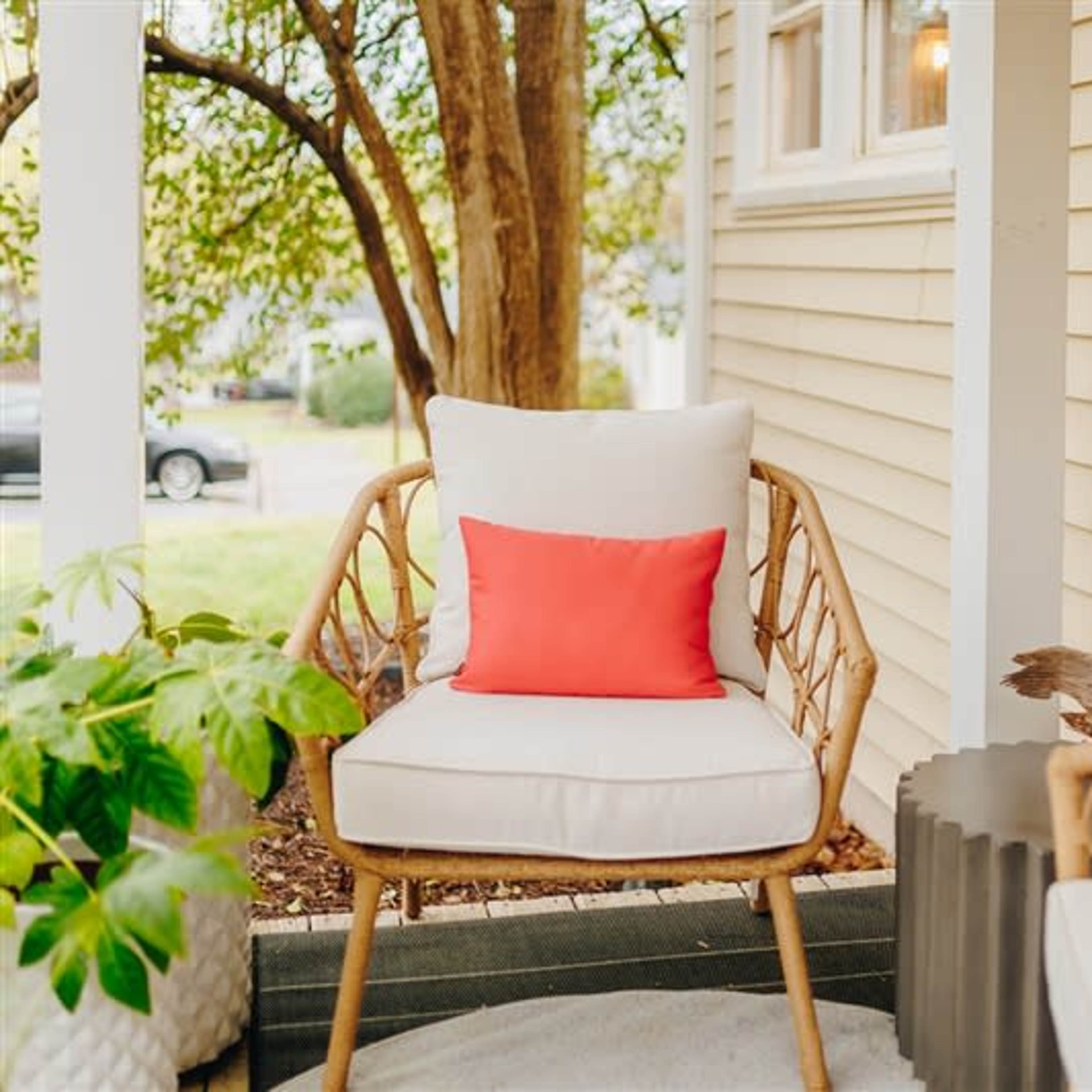 A rattan chair with a white cushion and a pink pillow is positioned on a porch alongside a small table and greenery.