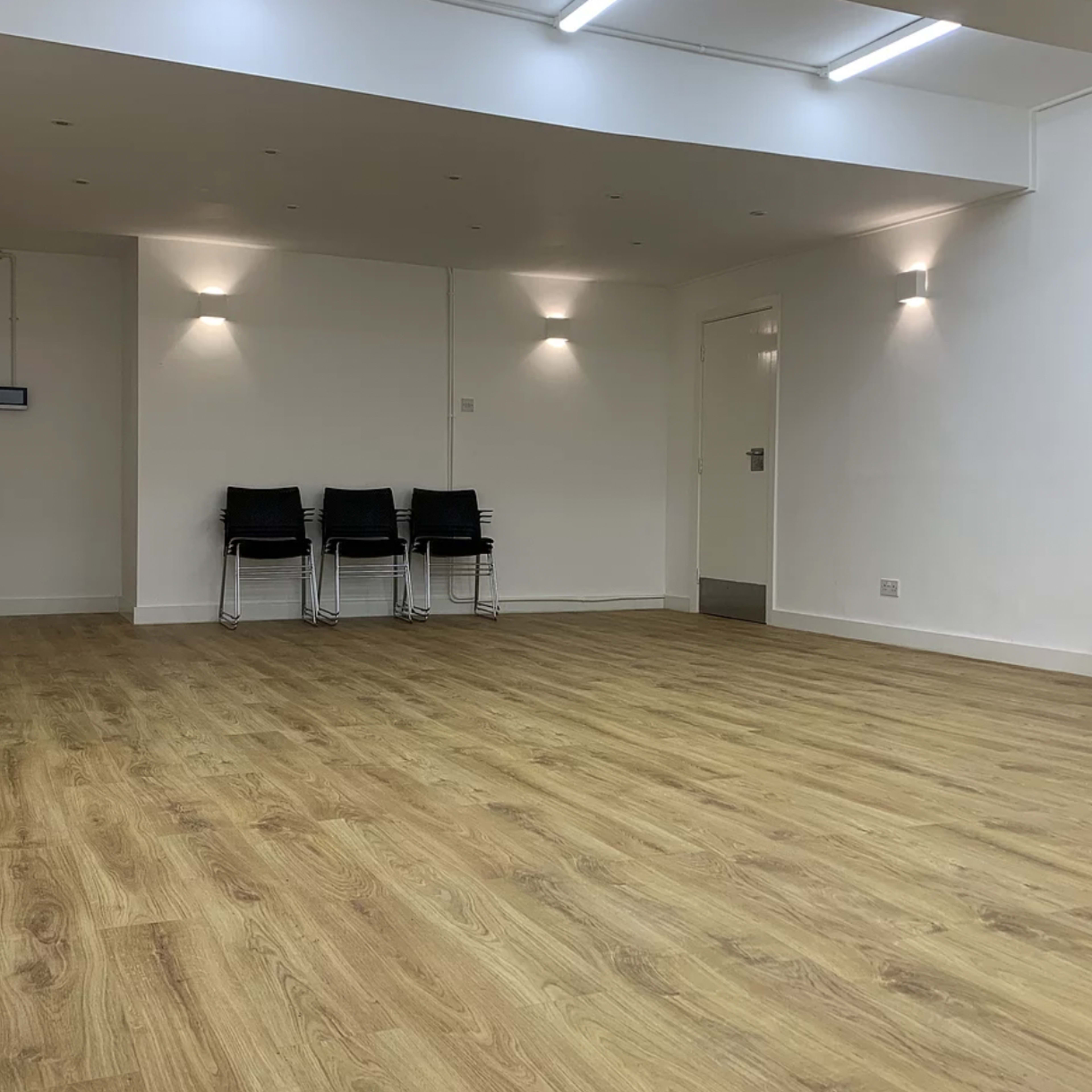 A spacious, empty room featuring wooden flooring, white walls, and a few black chairs lined against one wall.
