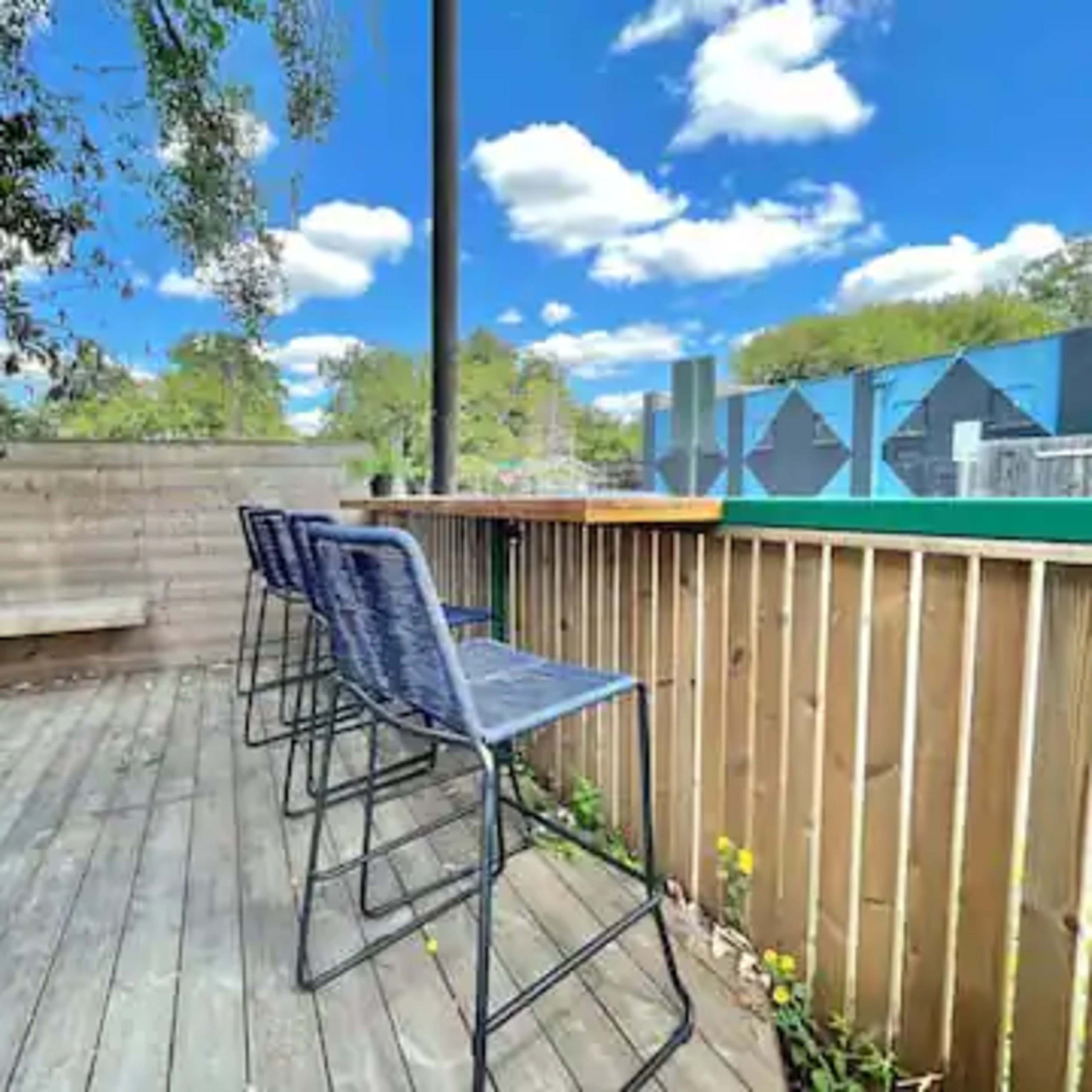 A wooden deck with three high stools facing a green area under a blue sky with clouds.