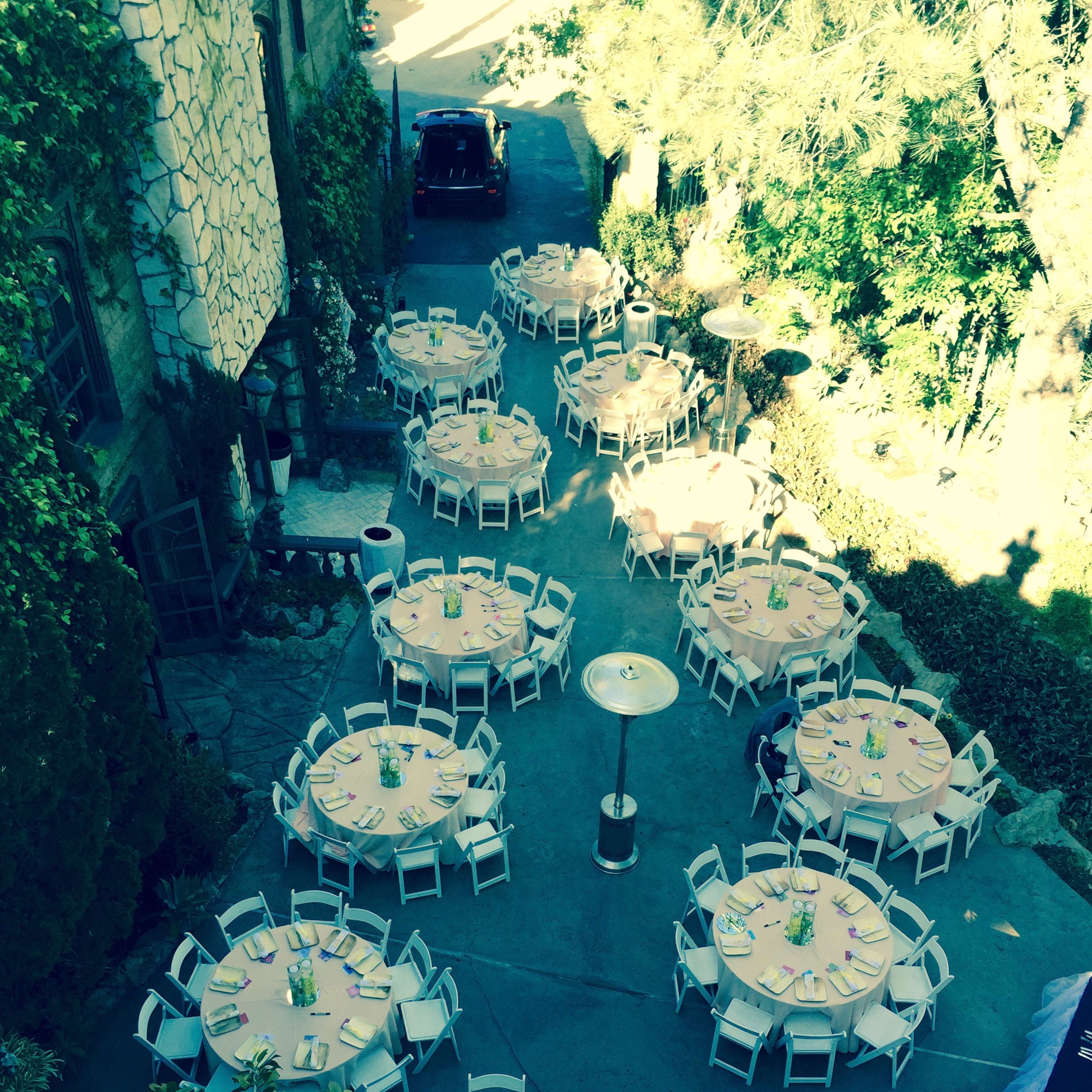 An outdoor event setup with multiple round tables arranged neatly in a courtyard, surrounded by greenery.