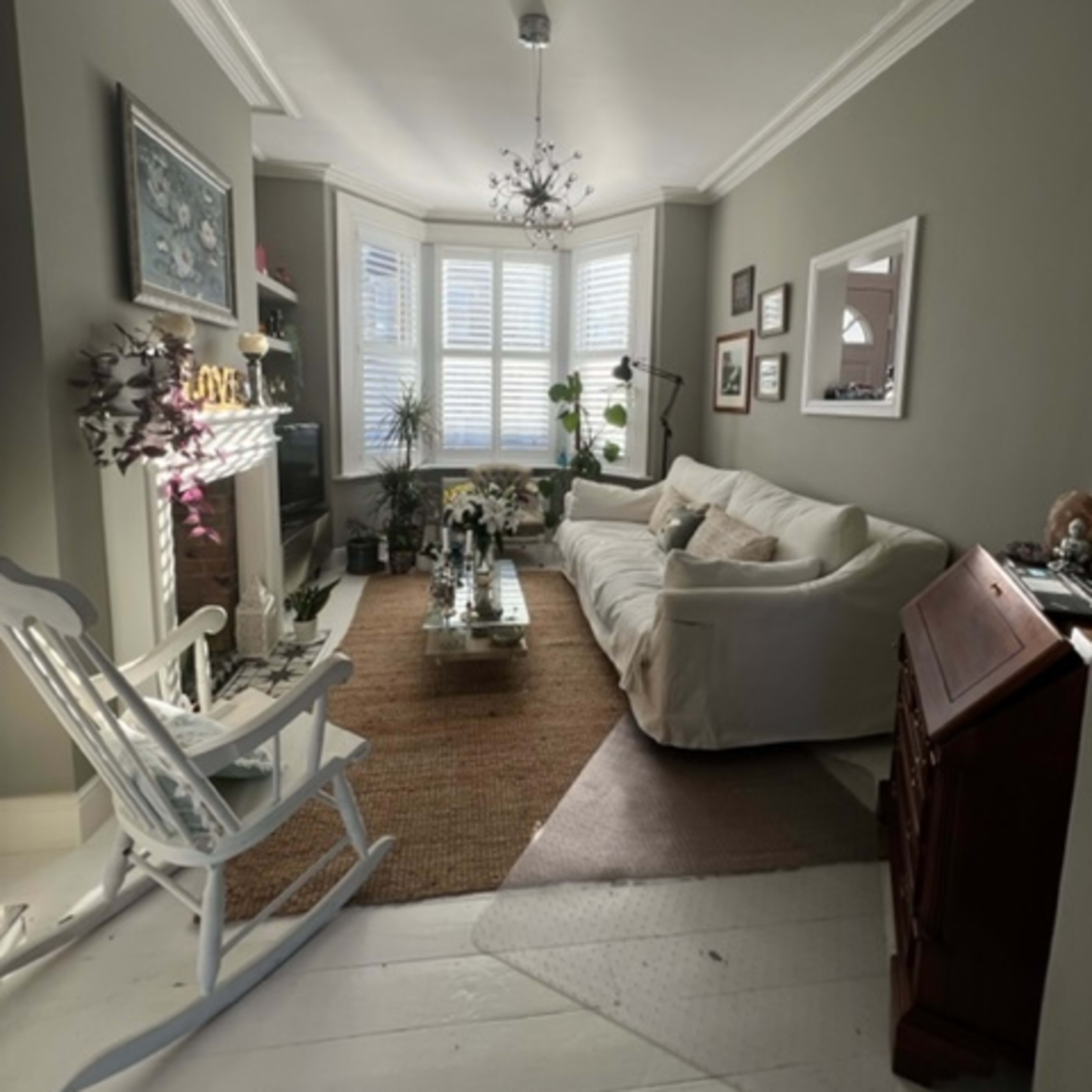 A living room featuring a light-colored sofa, a glass coffee table, a rocking chair, and a decorative fireplace, with natural light coming through bay windows.