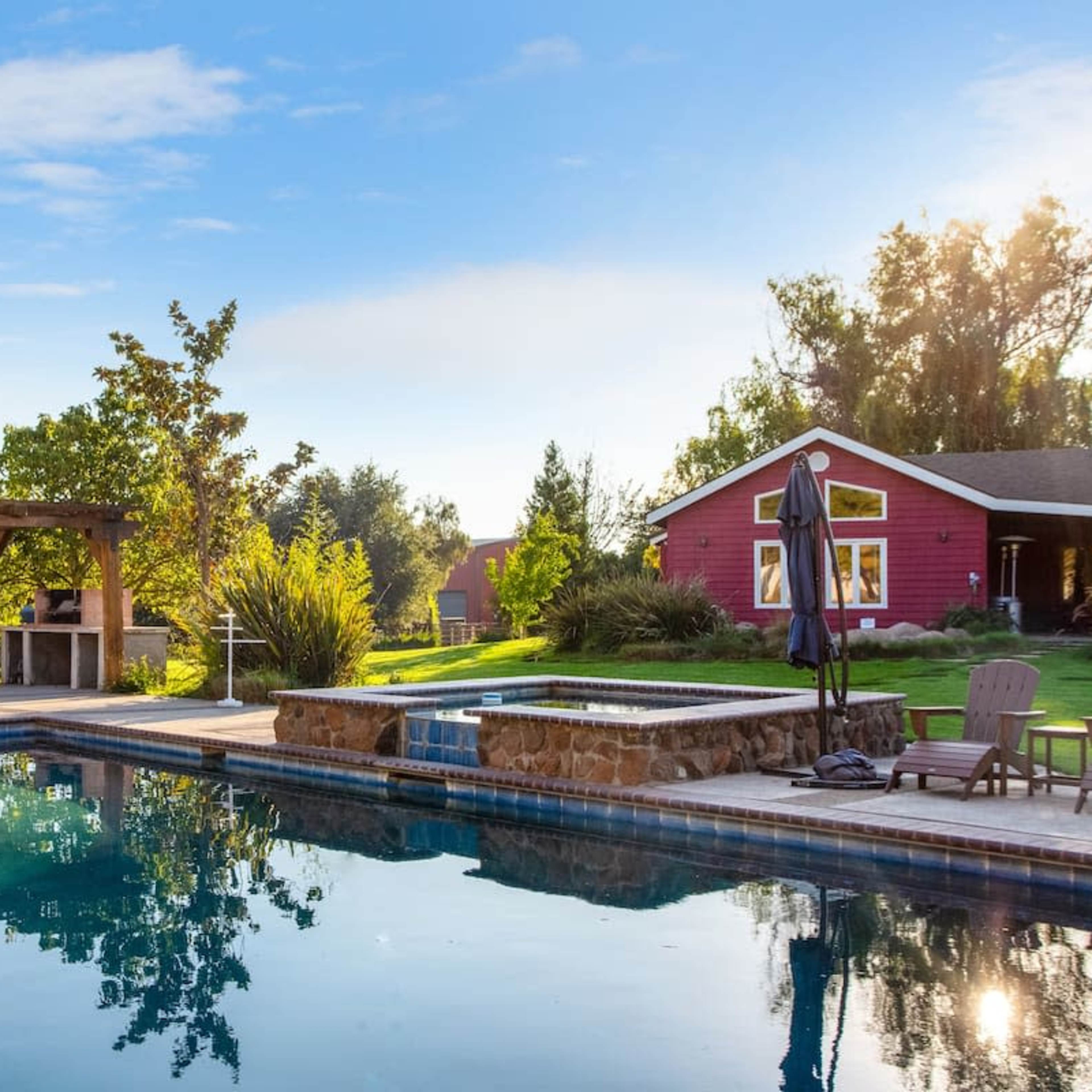 A landscaped backyard features a swimming pool, a wooden pergola with seating, and a red house surrounded by trees and grass.