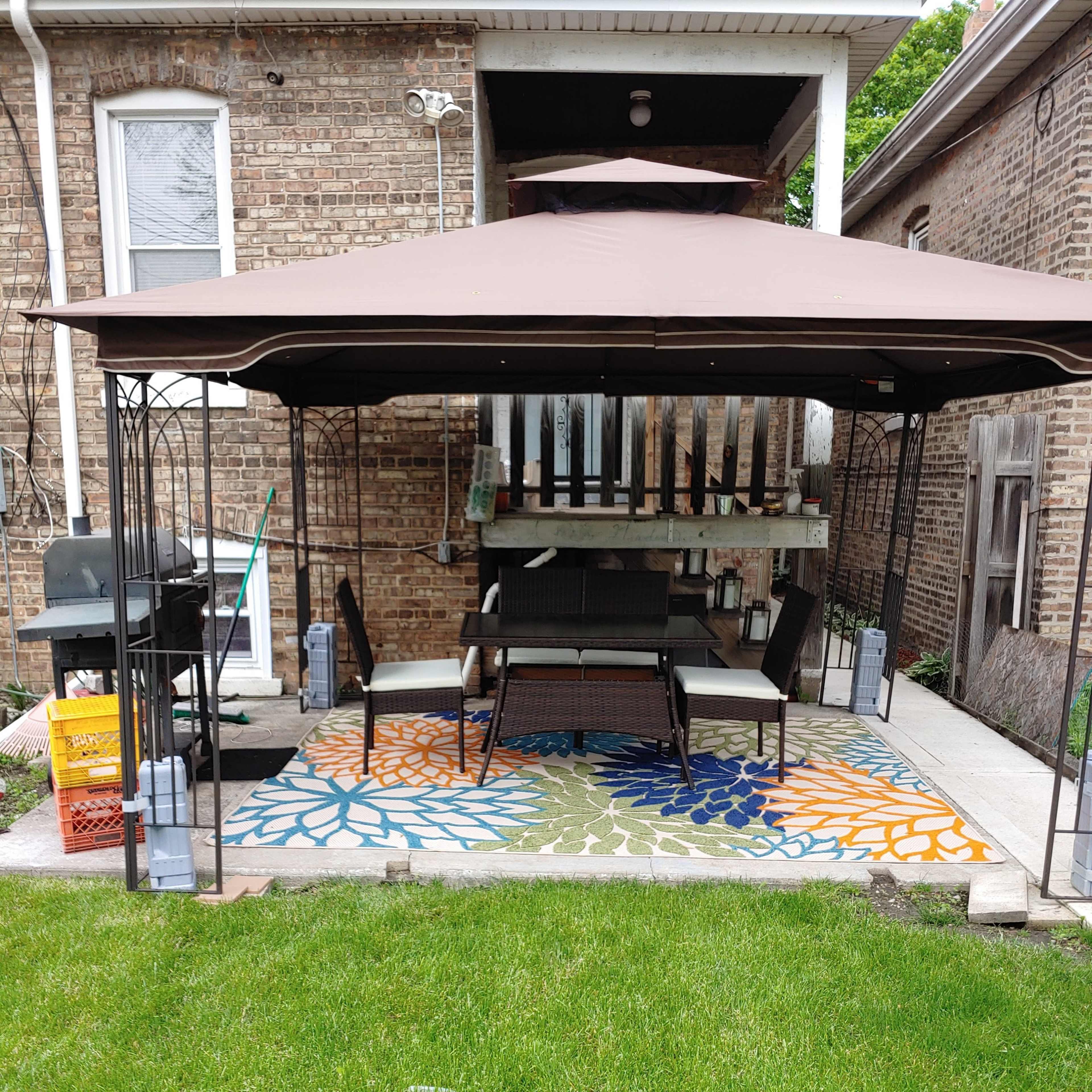 A metal gazebo with a brown canopy shelters a seating area on a patterned rug in a backyard with grass and nearby cooking equipment.