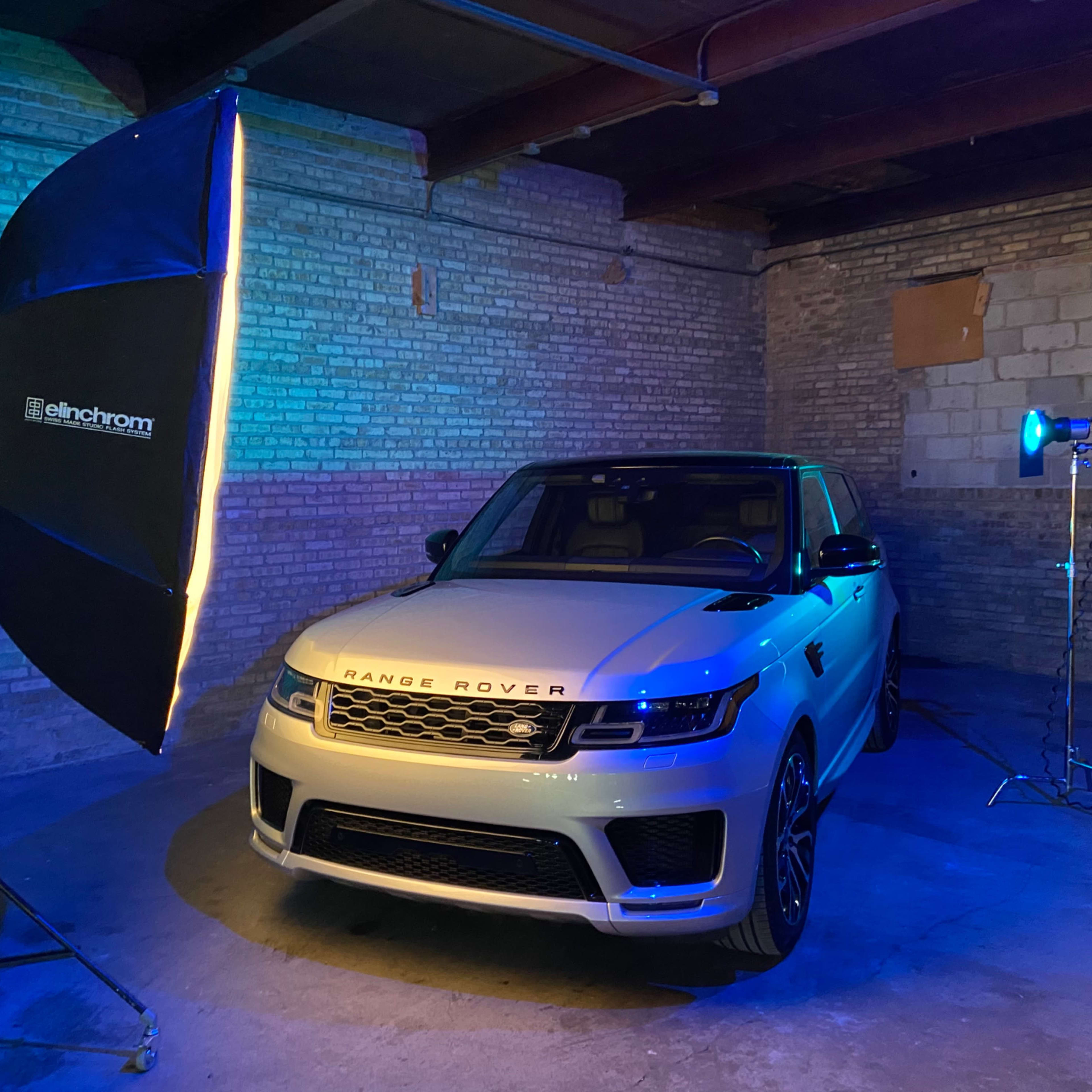 A white Range Rover is parked in a dimly lit industrial space, illuminated by a large softbox and a spotlight.