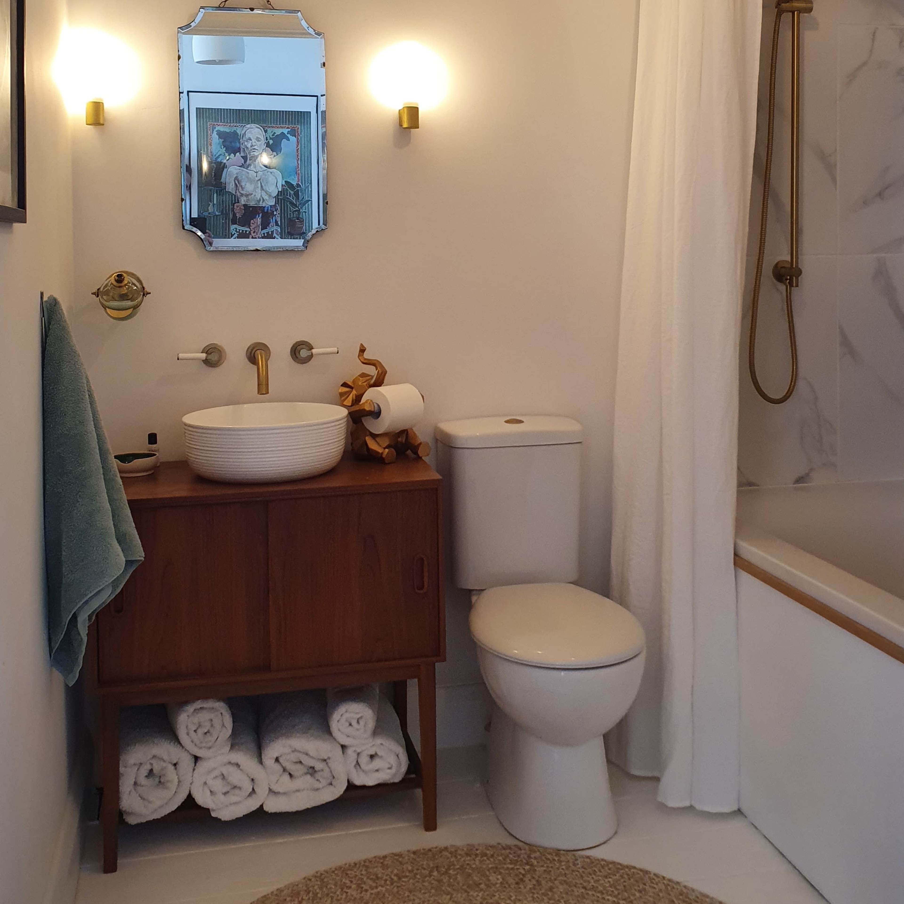 A bathroom featuring a wooden vanity with a round sink, a toilet, a shower with a gold showerhead, white curtains, and neatly stacked towels.
