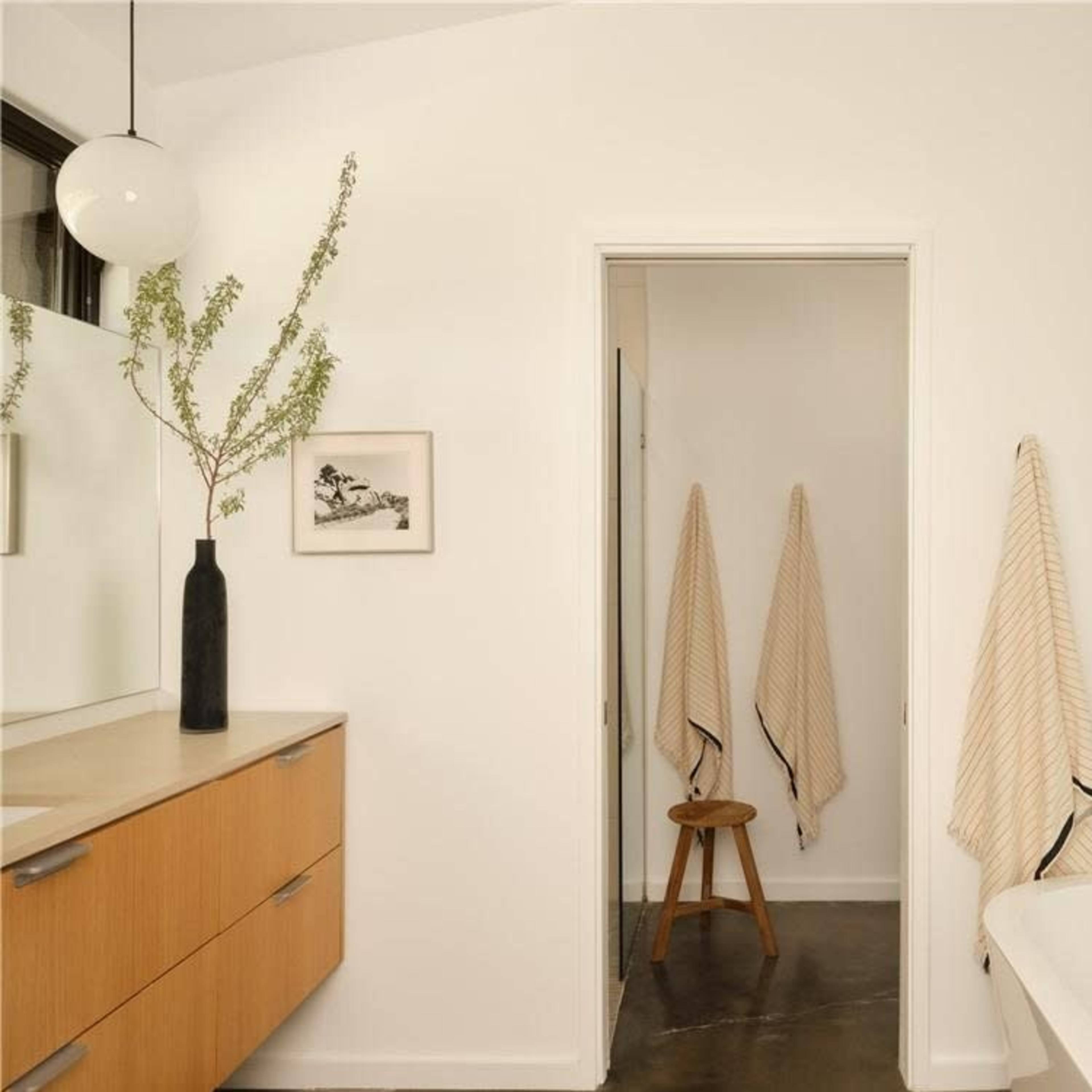A modern bathroom features a wooden vanity, a potted plant, three hanging towels, and a stool next to a freestanding bathtub.