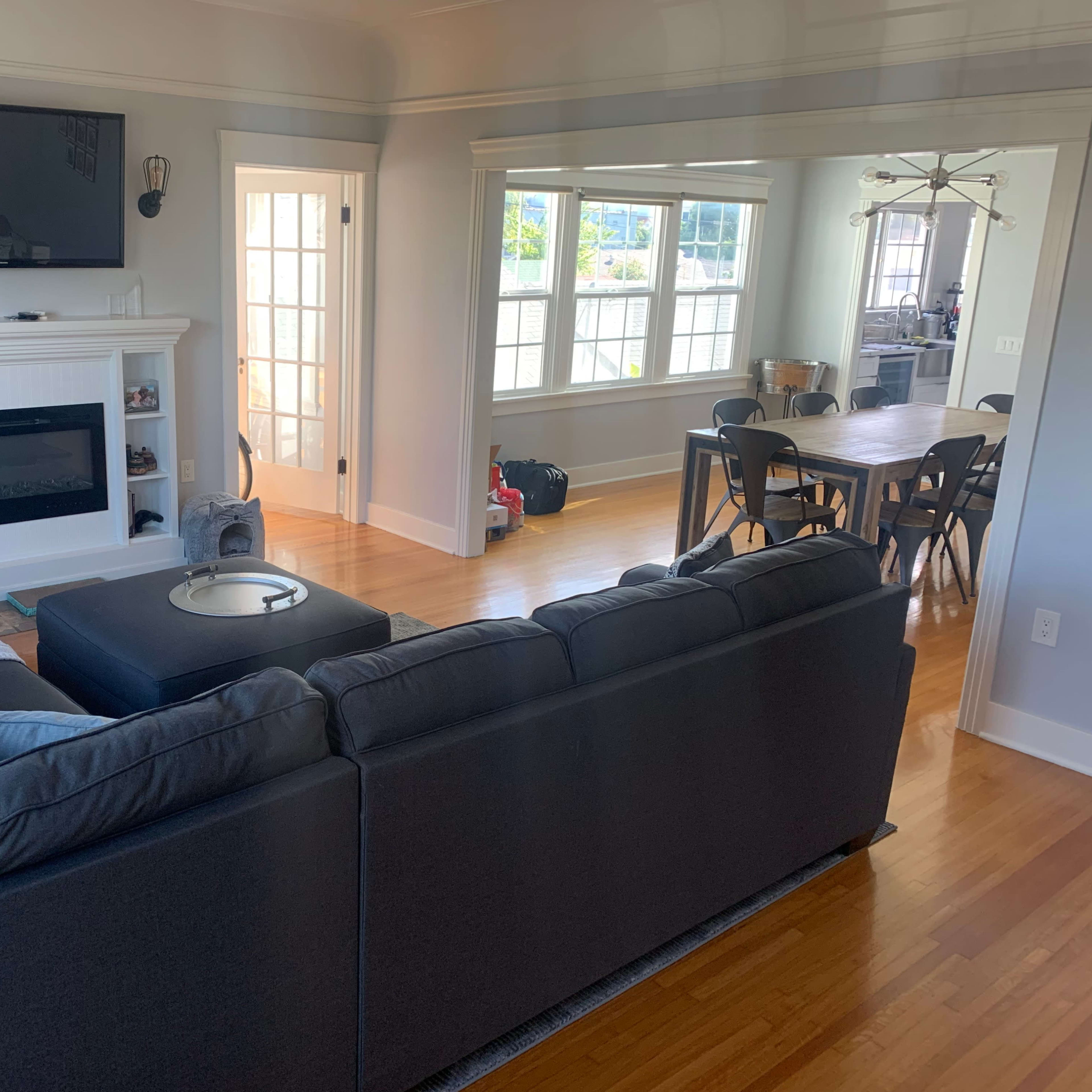 A living room with a gray sectional couch, a wall-mounted television above a white fireplace, and a dining area visible through an open doorway.