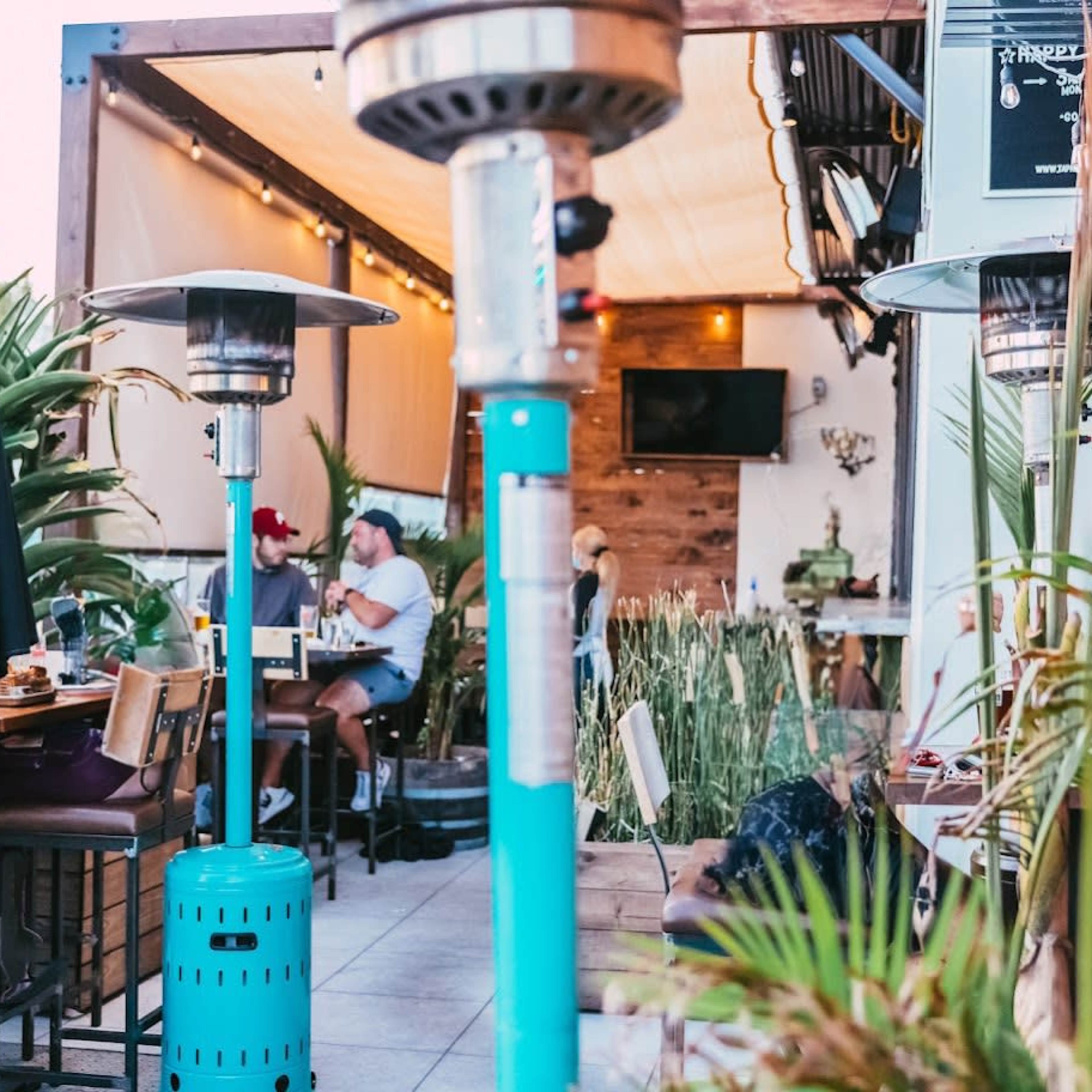 A dining area with outdoor heaters features tables, plants, and patrons engaged in conversation.